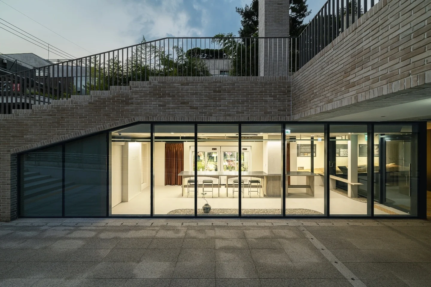 Modern building with large glass windows showing an interior kitchen and seating area, brick exterior, outdoor staircase on the left, and a terrace with a metal railing on upper level.