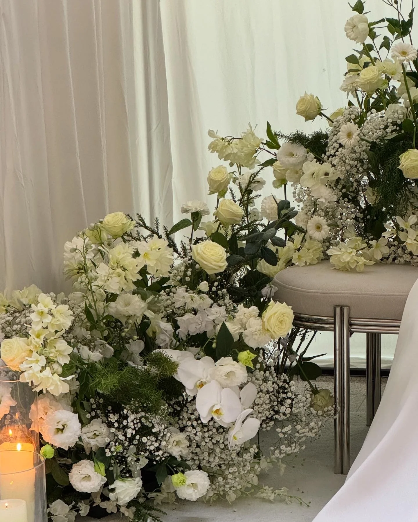 Elegant floral arrangement with white and cream flowers, including orchids, roses, and baby's breath, near a white curtain and a beige cushioned stool.