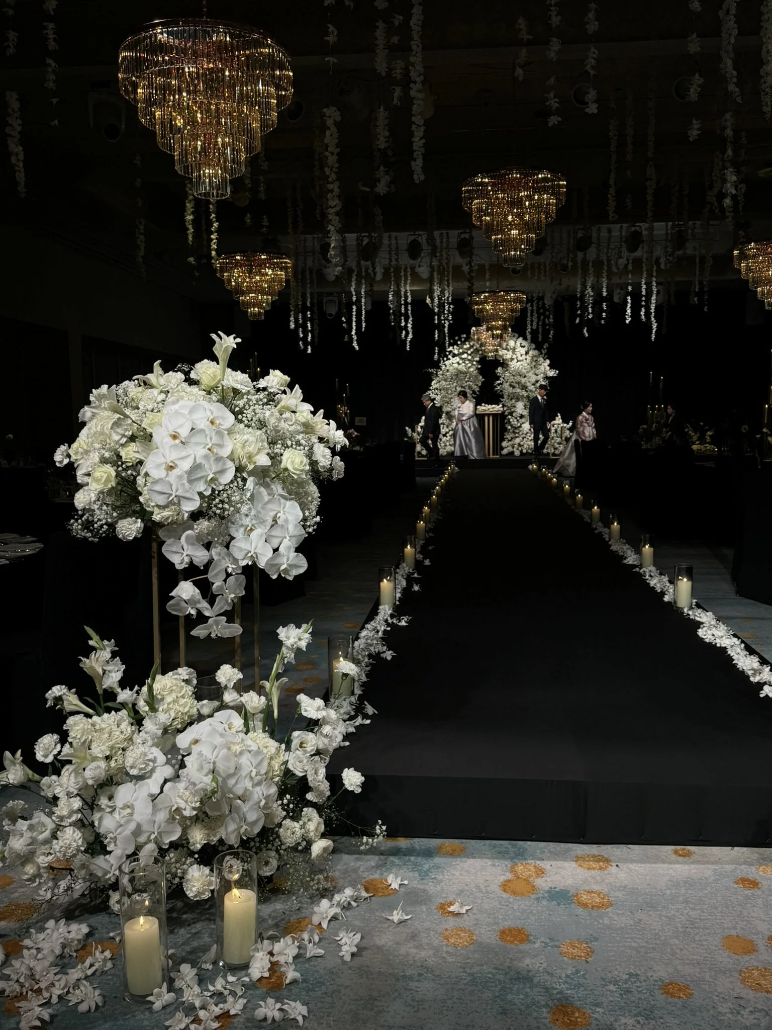 A dimly lit wedding aisle decorated with white flowers and candles, with a floral arch at the front and chandeliers hanging from the ceiling.