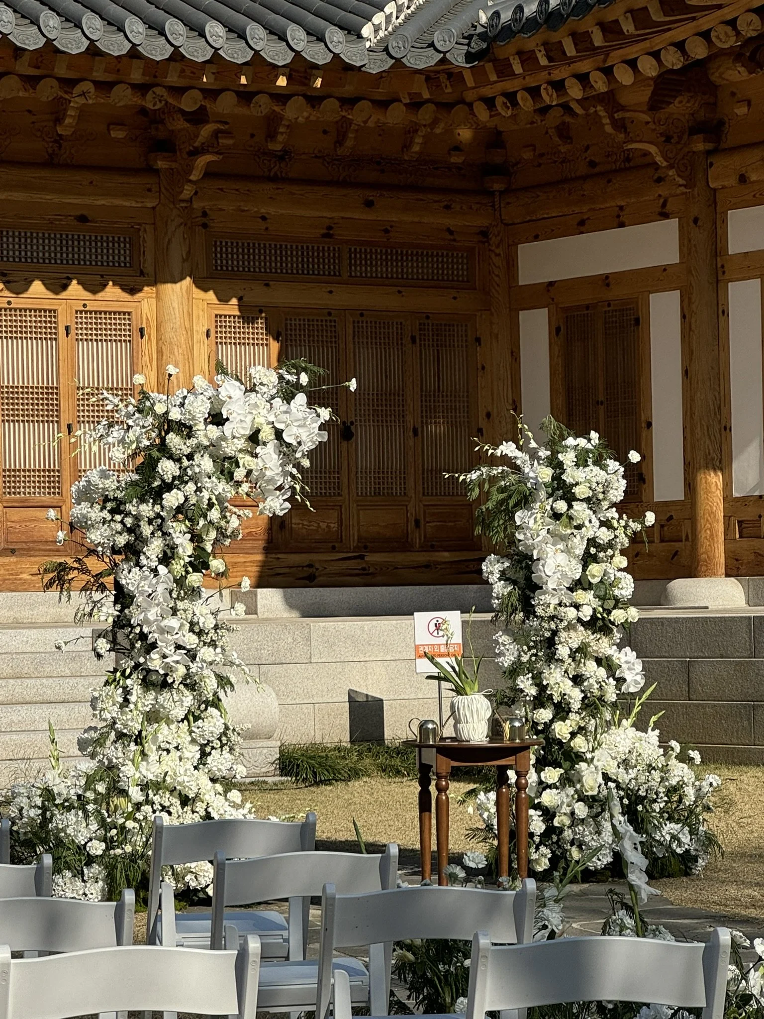 A wedding altar decorated with white flowers and greenery in front of a traditional wooden building with a tiled roof. White chairs are arranged facing the altar, and there is a small table with a vase and a sign.