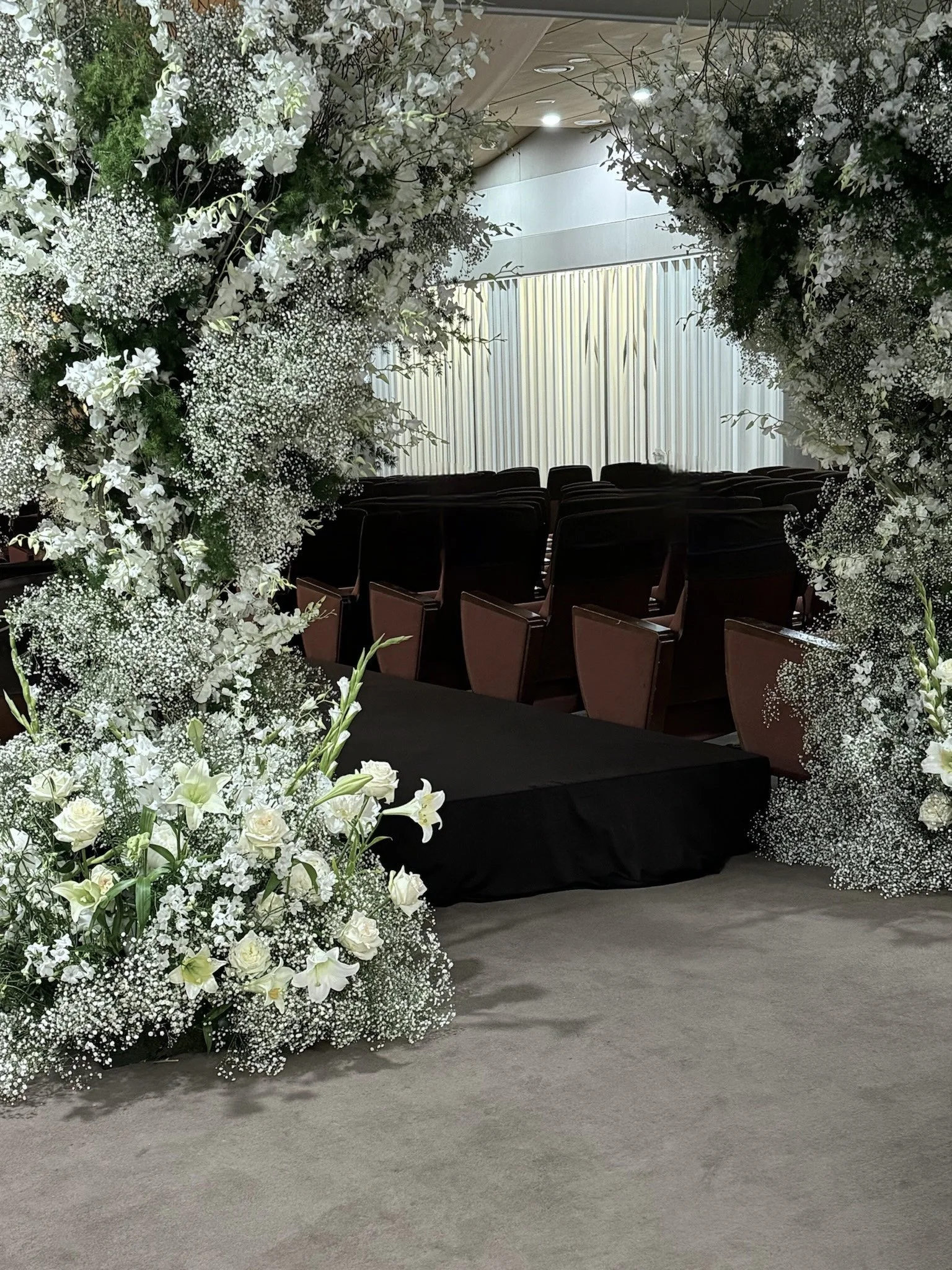 A floral wedding arch with white flowers and greenery, with rows of chairs and a stage in the background.