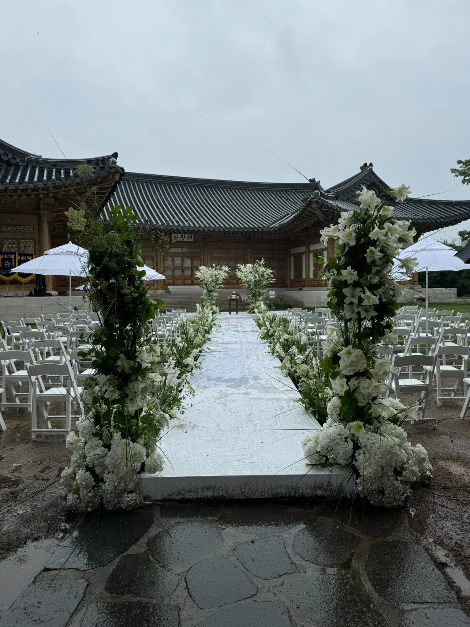 Wedding altar decorated with white flowers and greenery, set outdoors on a rainy day with traditional Korean architecture in the background.