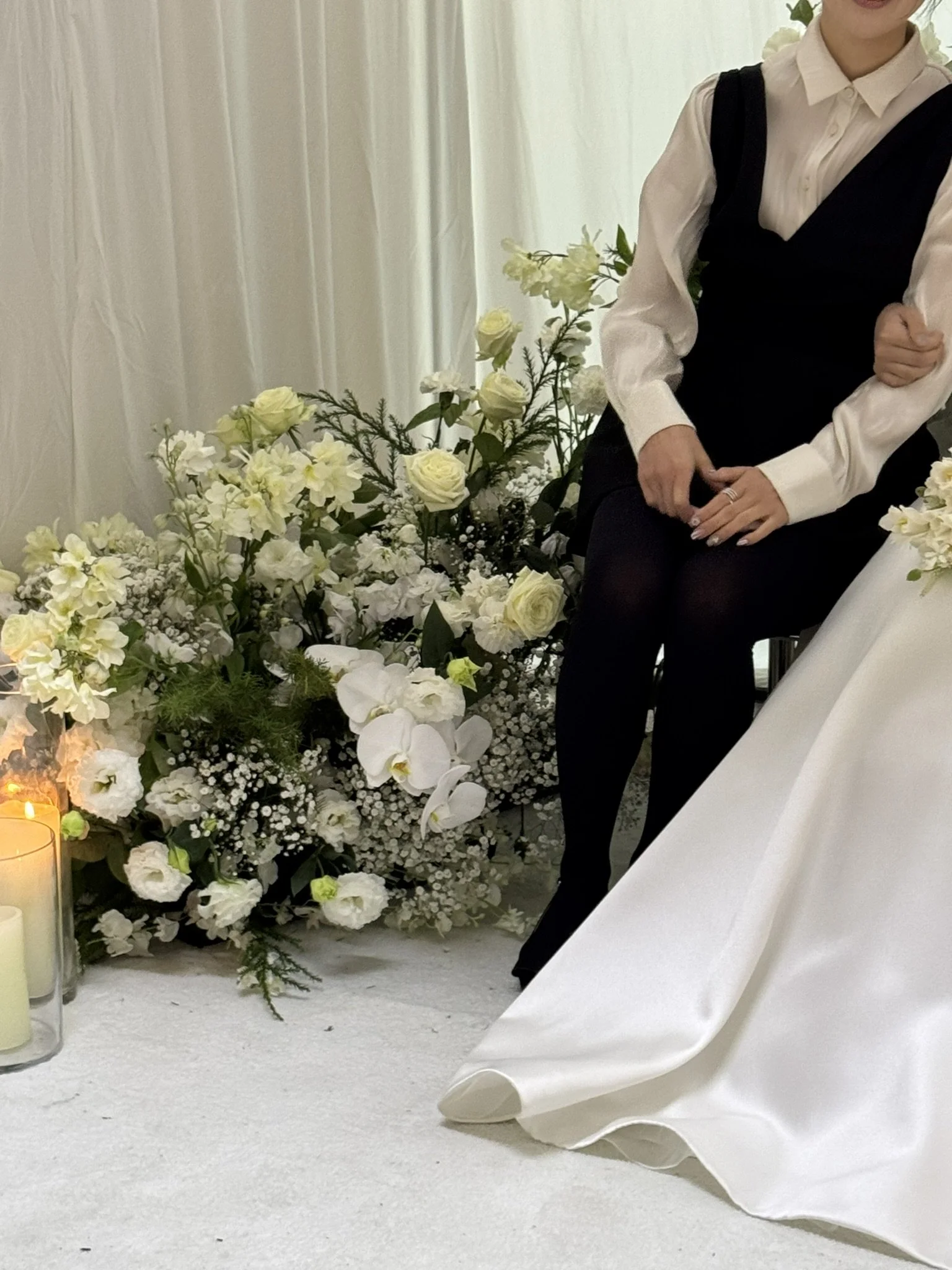 A woman in a white blouse and a black dress sitting beside a large white floral arrangement with candles on the floor.
