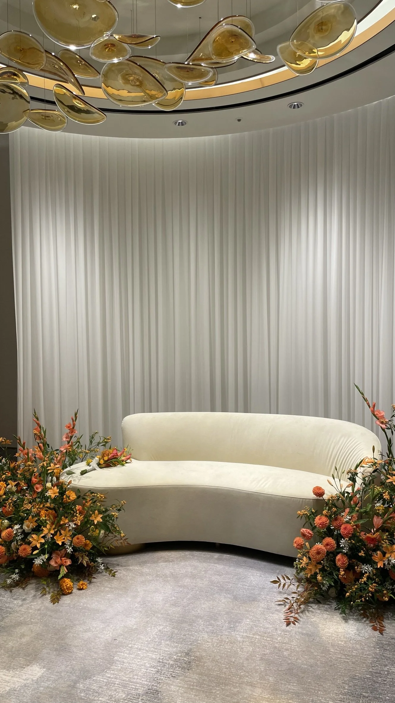 An elegant room with a curved cream-colored sofa, floral arrangements on both sides, a white curtain backdrop, and an overhead modern gold and glass chandelier.