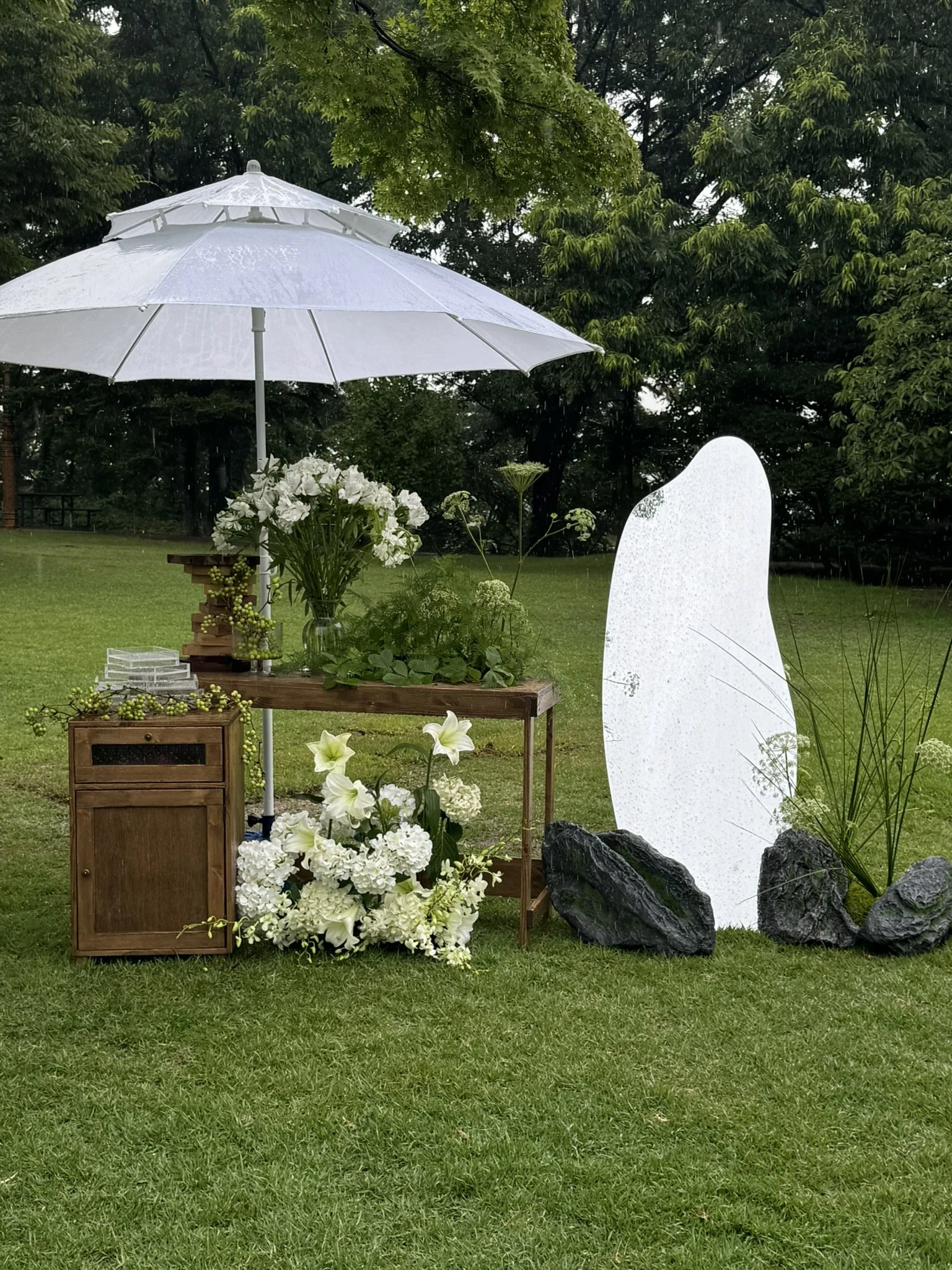 A floral display outdoors with white flowers, green foliage, a large white umbrella, a small wooden table, and decorative rocks and a white stone sculpture in a grassy area with trees in the background.