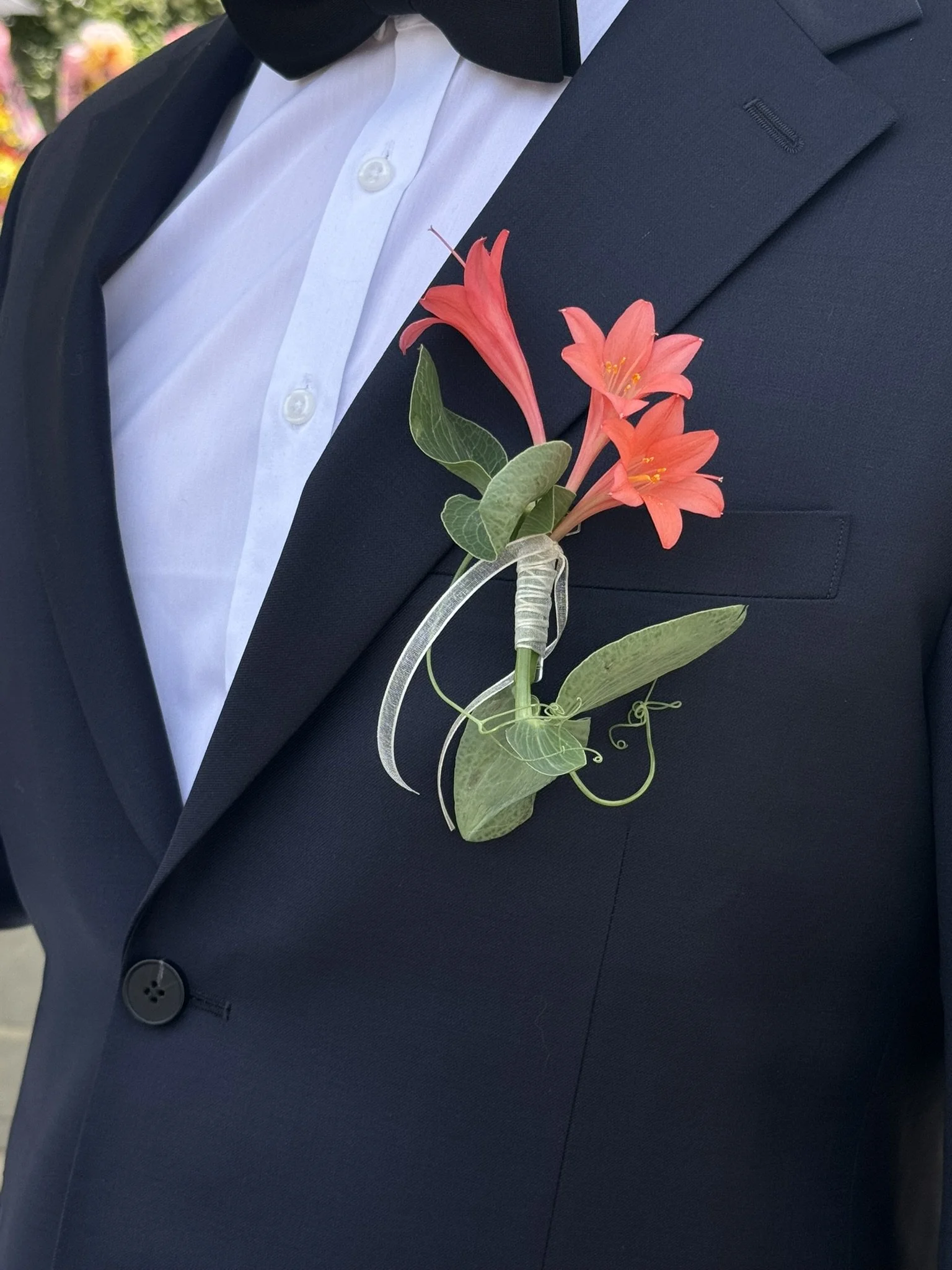 Close-up of a person wearing a black tuxedo with a white shirt and a black bow tie, sporting a pink floral boutonniere pinned to the left lapel.