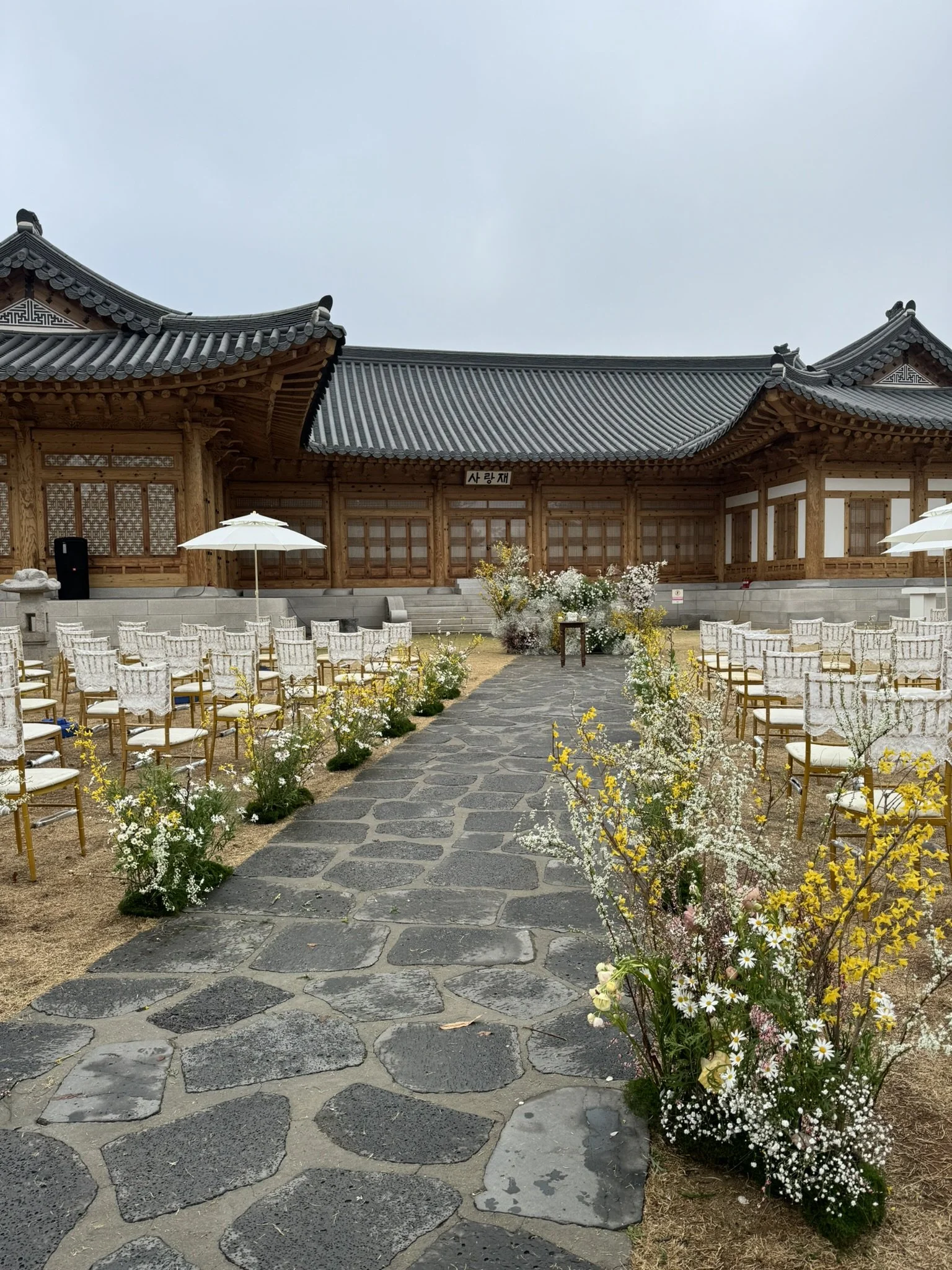 Outdoor venue with traditional Korean style wooden building, stone pathway, white chairs, umbrellas, and floral decorations, set for an event or ceremony.