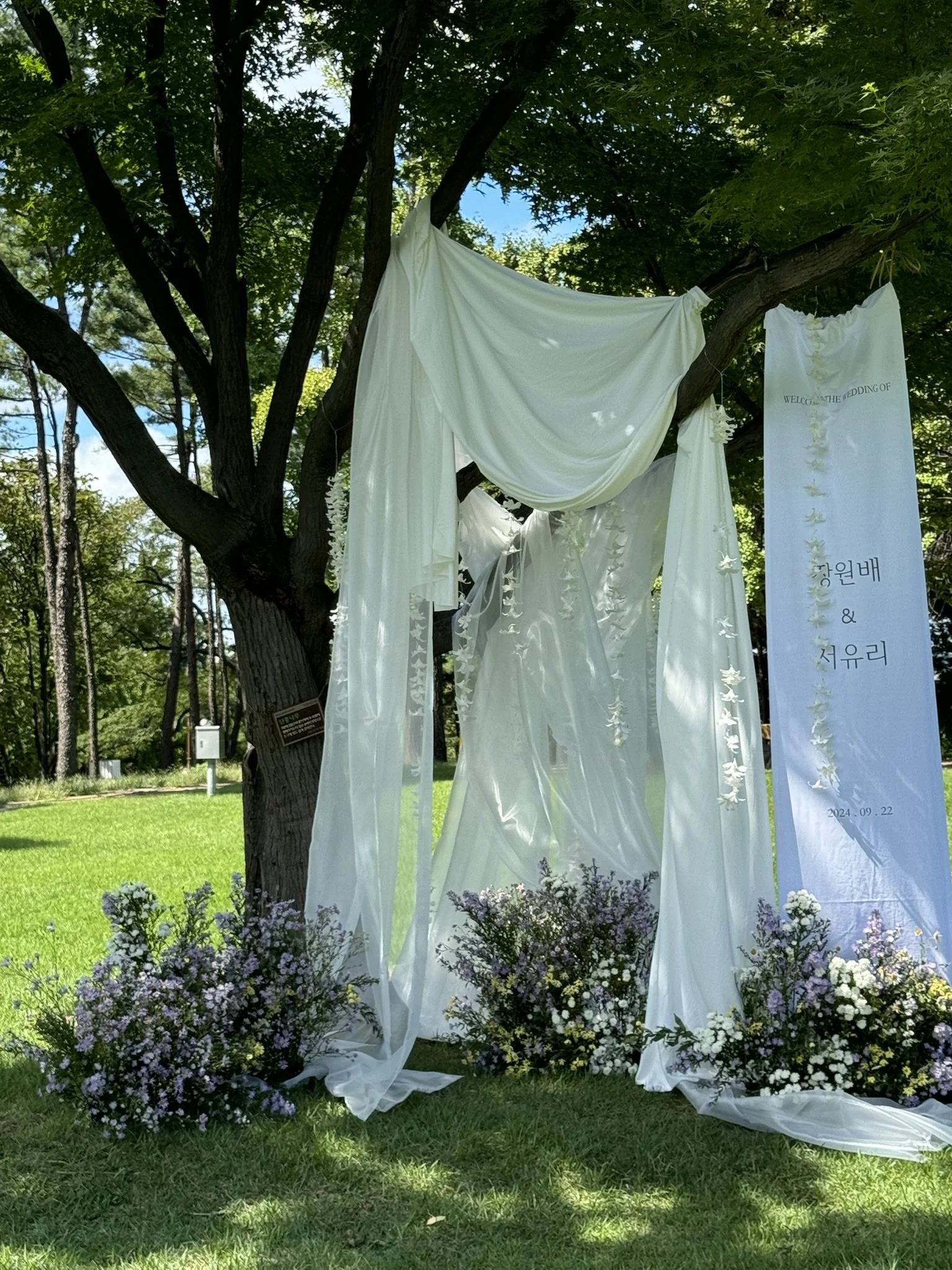 Outdoor wedding ceremony setup with white drapery and floral arrangements under a tree.