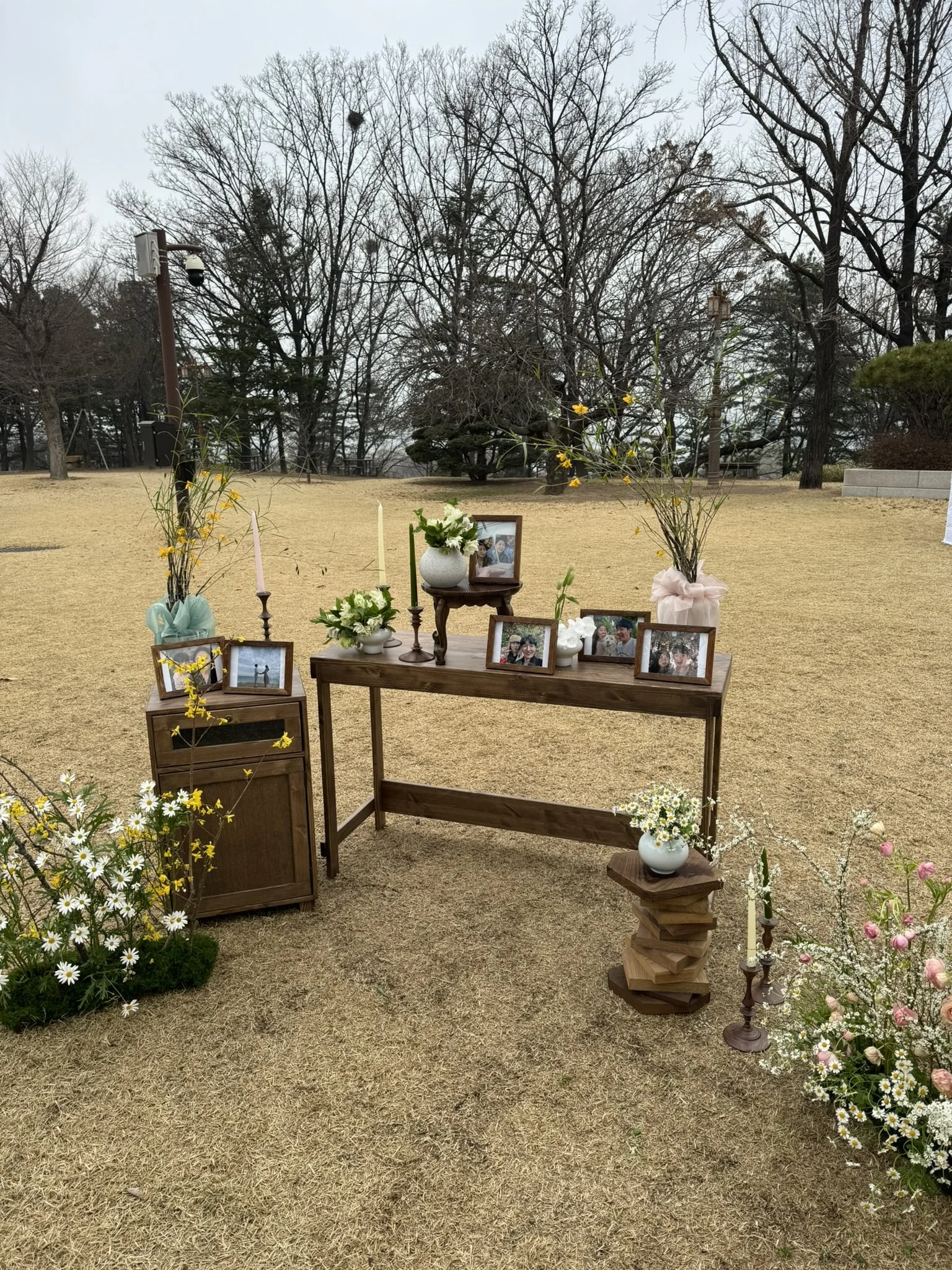 An outdoor memorial setup with wooden tables and shelves, decorated with framed photographs, vases of flowers, and candles, surrounded by bushes and trees.