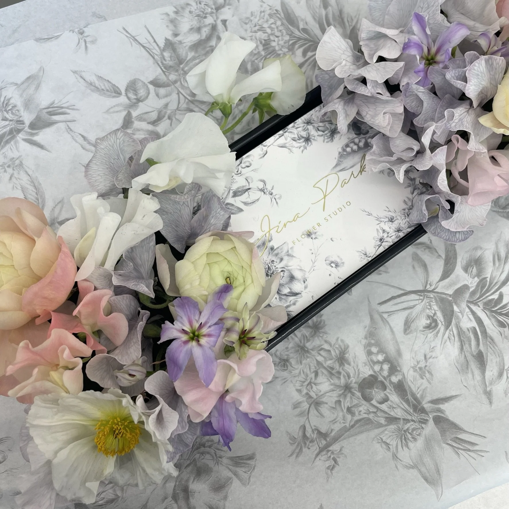A floral arrangement with pastel-colored flowers including pink, purple, white, and green, placed on a gray floral patterned paper with a white card that has gold cursive writing reading 'Jina Park' and 'Flower Studio'.