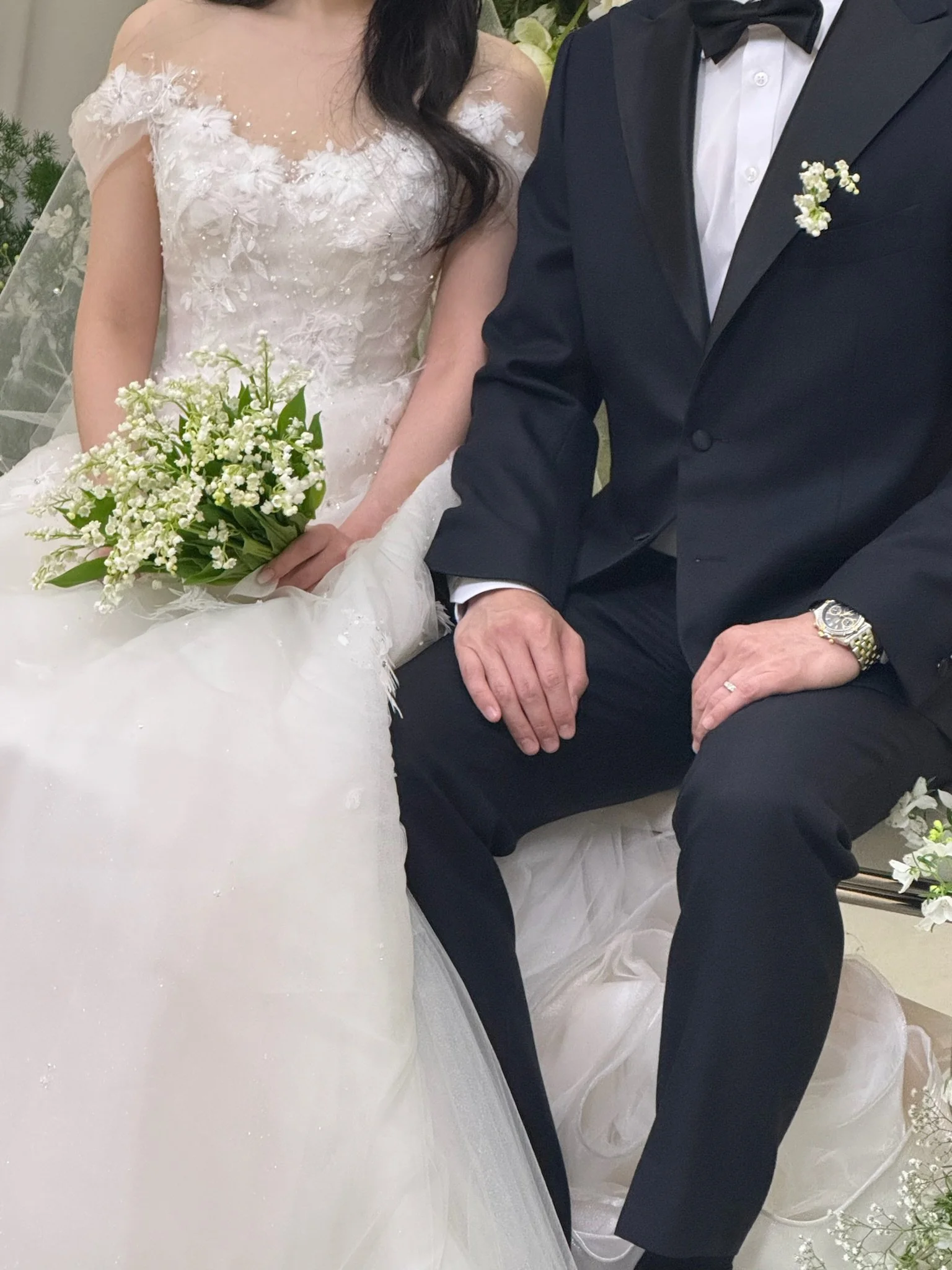 A bride in a white wedding gown holding a bouquet, seated next to a groom in a black tuxedo with a white shirt and black bow tie, with flowers and greenery in the background.
