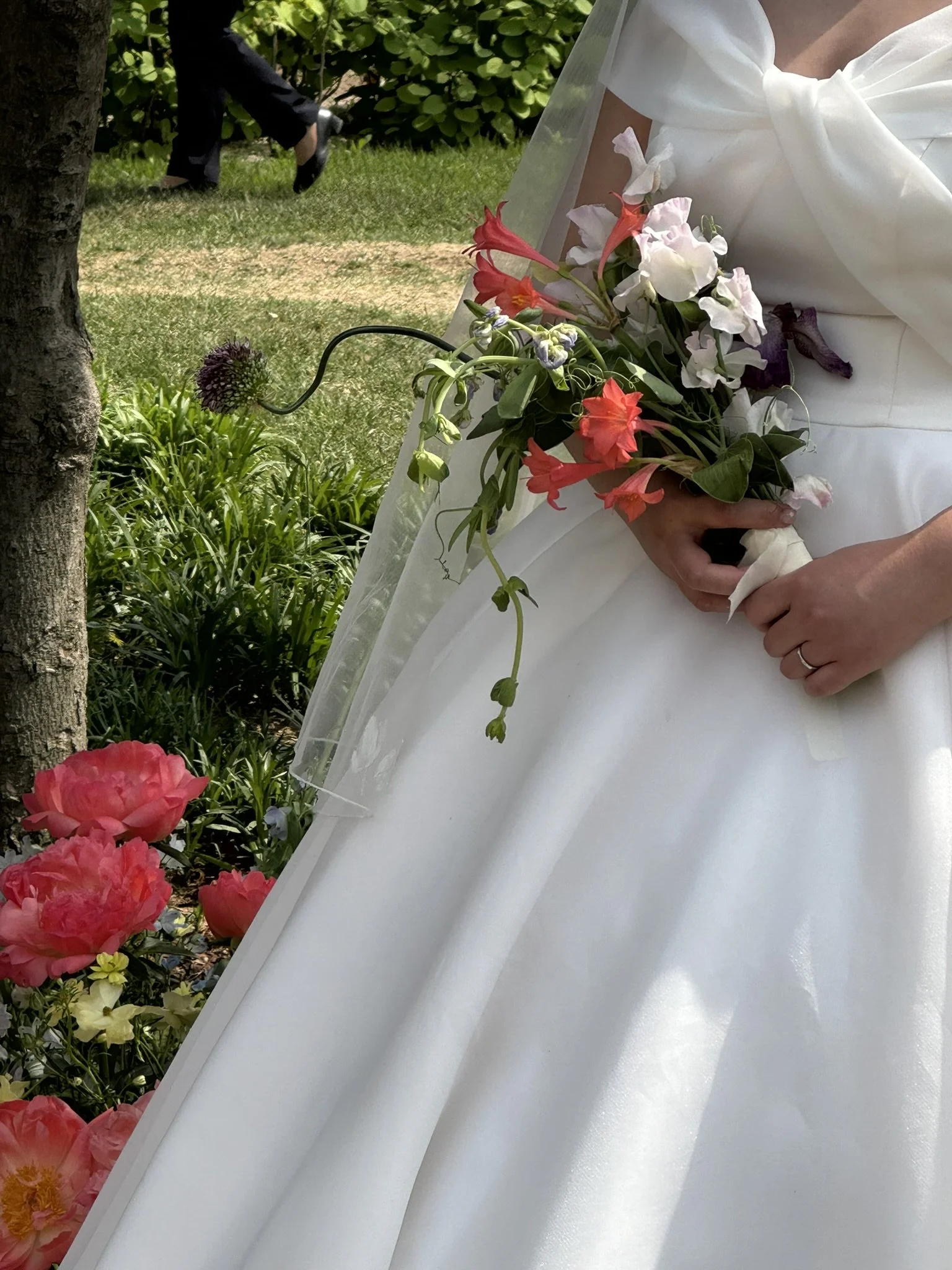 A bride wearing a white wedding dress holds a bouquet of colorful flowers, including pink, white, and purple blooms, outdoors near a tree and green bushes.
