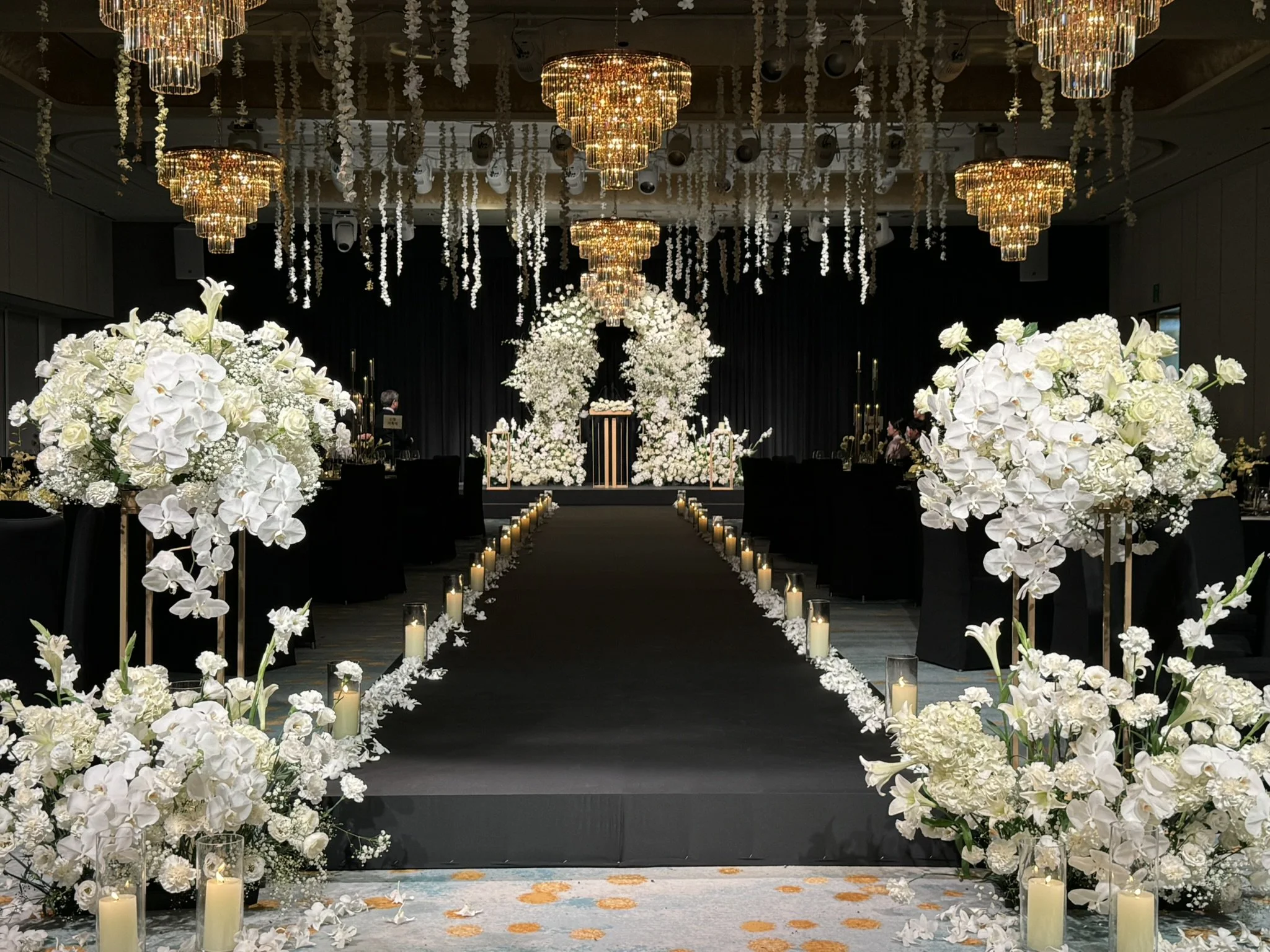 Elegant wedding ceremony setup with a black aisle runner, white floral arrangements, candles, and chandeliers in a grand hall.