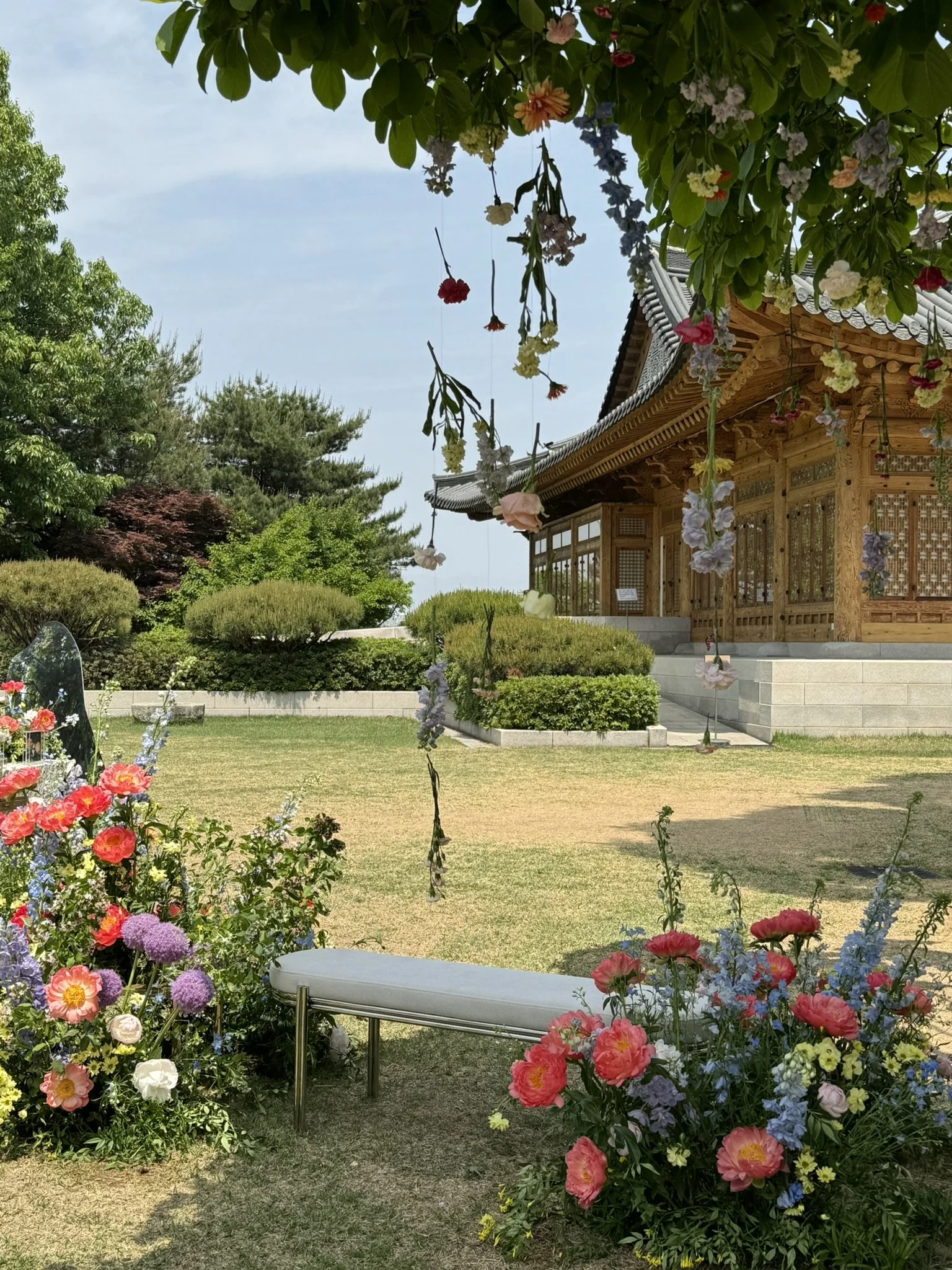 A traditional wooden Asian-style building with ornate roof details, surrounded by a garden with various colorful flowers, green bushes, and trees. There are hanging flower decorations and a white bench in the garden.