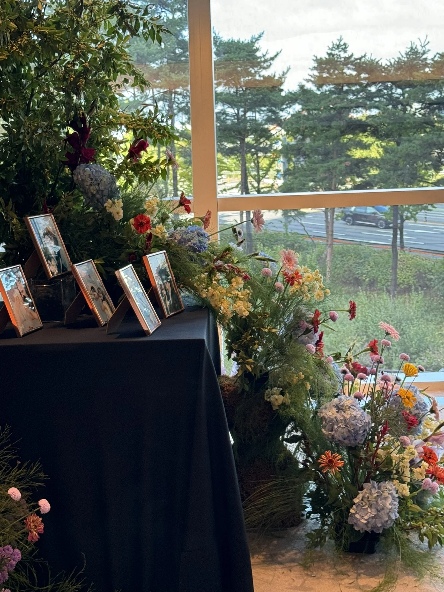 Indoor display of framed photographs and colorful flower arrangements near a large window with trees and a highway outside.