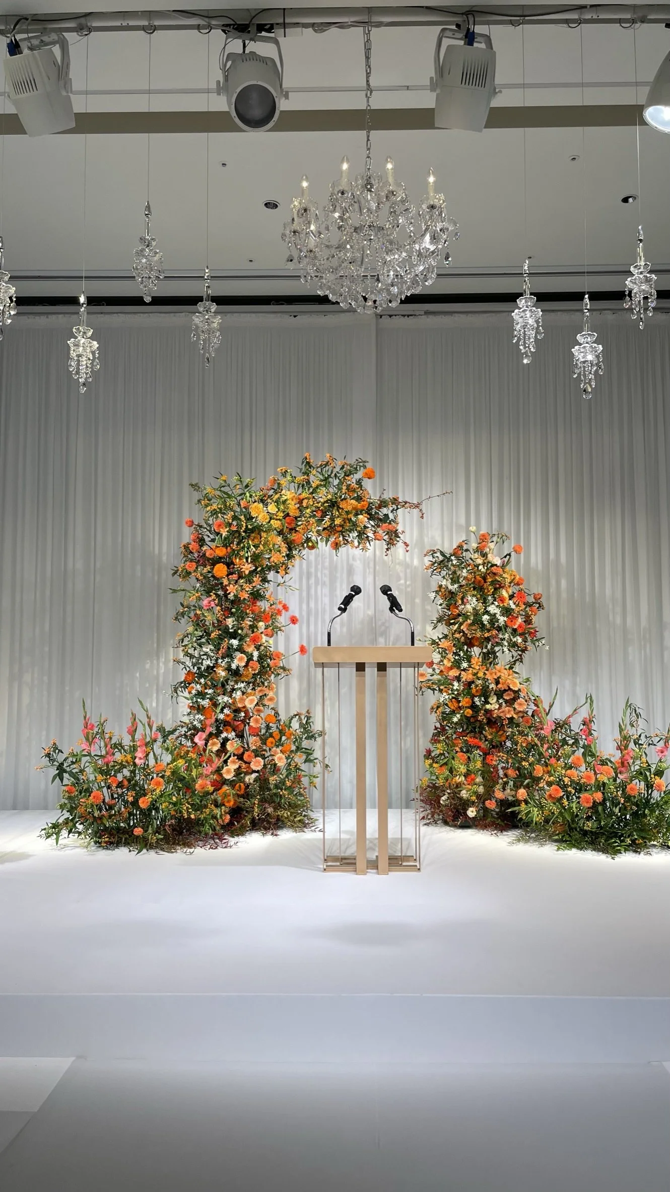 A decorated stage with a wooden podium, colorful floral arrangements, hanging chandeliers, and ceiling lights, set for a wedding or event.