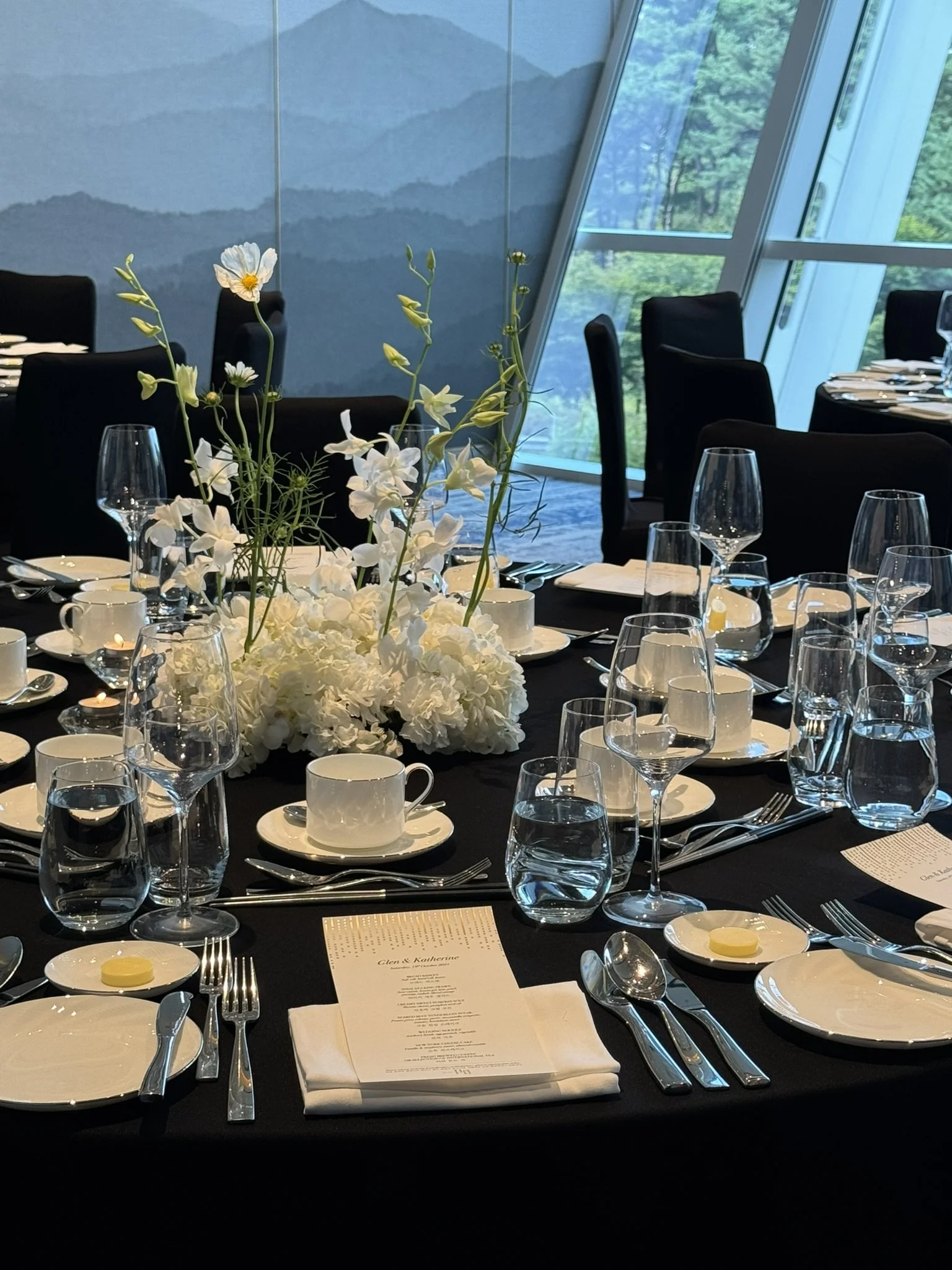 Elegant dining table set for a formal event with a black tablecloth, white plates, cups, silverware, and a centerpiece of white flowers, including roses and orchids, in a modern room with large windows and mountain views.