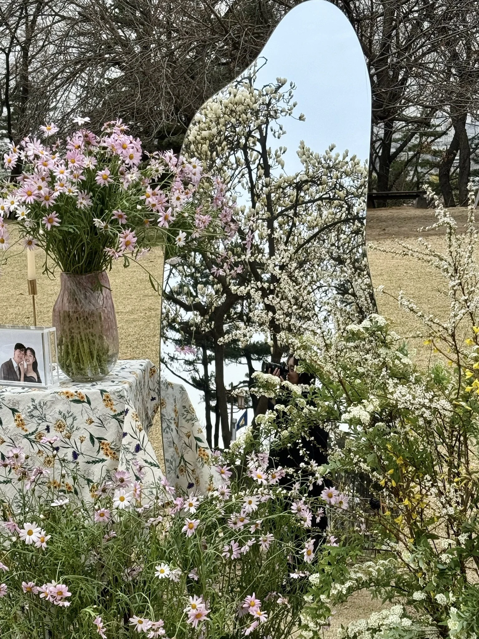 Flowers, a mirror reflecting trees, and a photograph on a table in an outdoor setting.