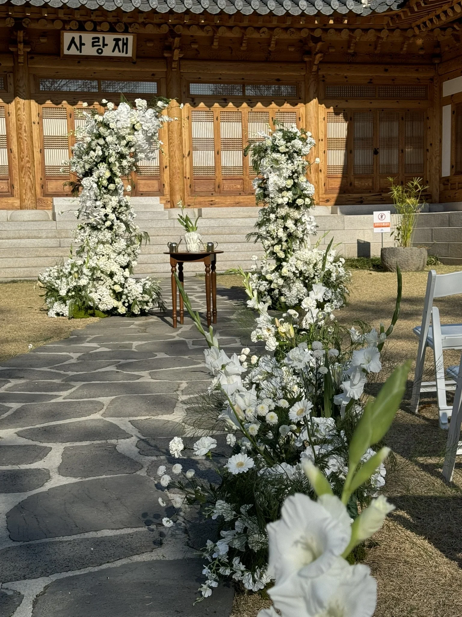 Decorated outdoor wedding aisle with white flowers, leading to a wooden building with Korean signs, on a stone pathway.
