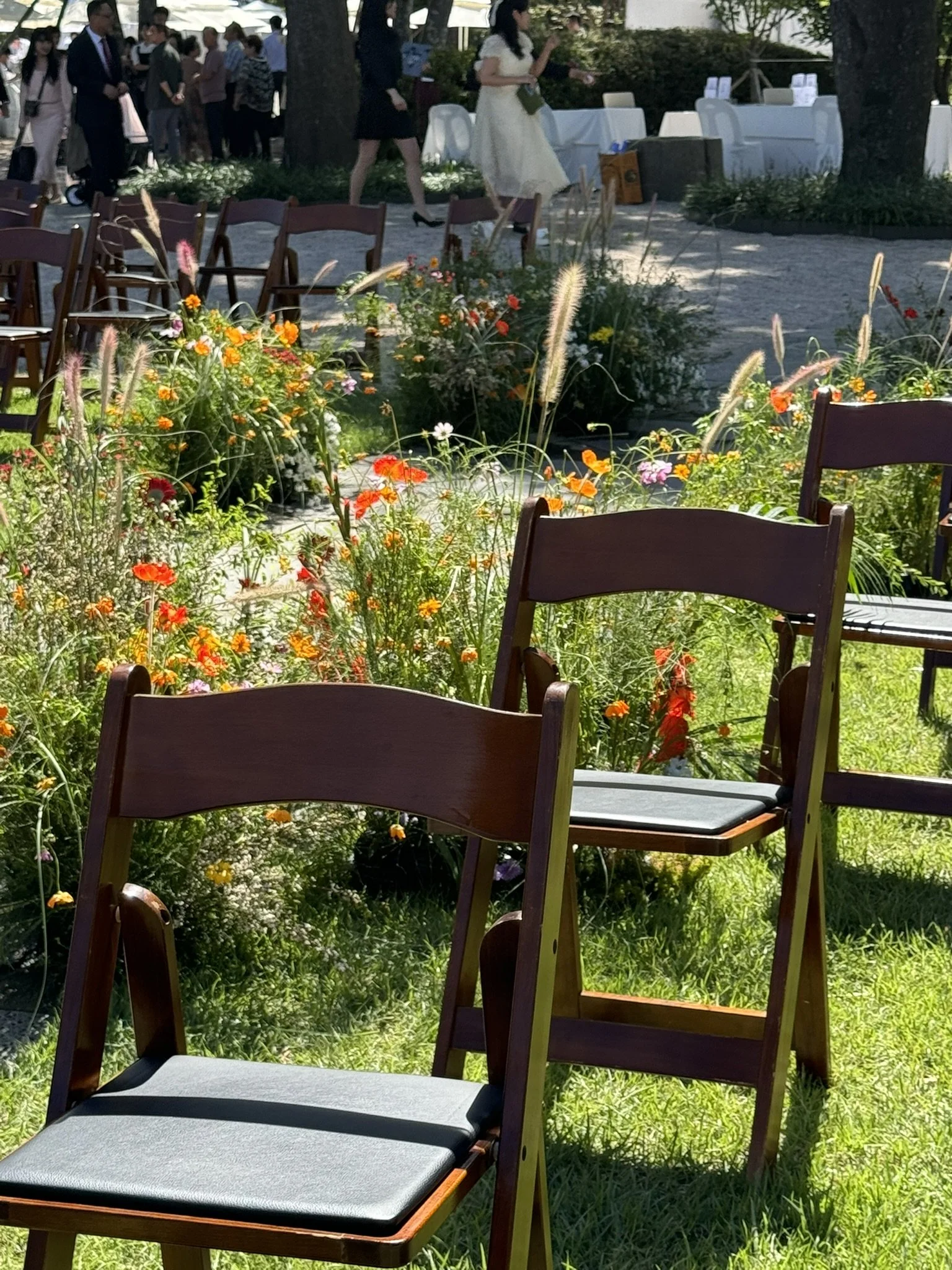 Empty wooden chairs with gray cushions arranged outdoors near a flower garden, with some people walking and standing in the background under a shaded area.