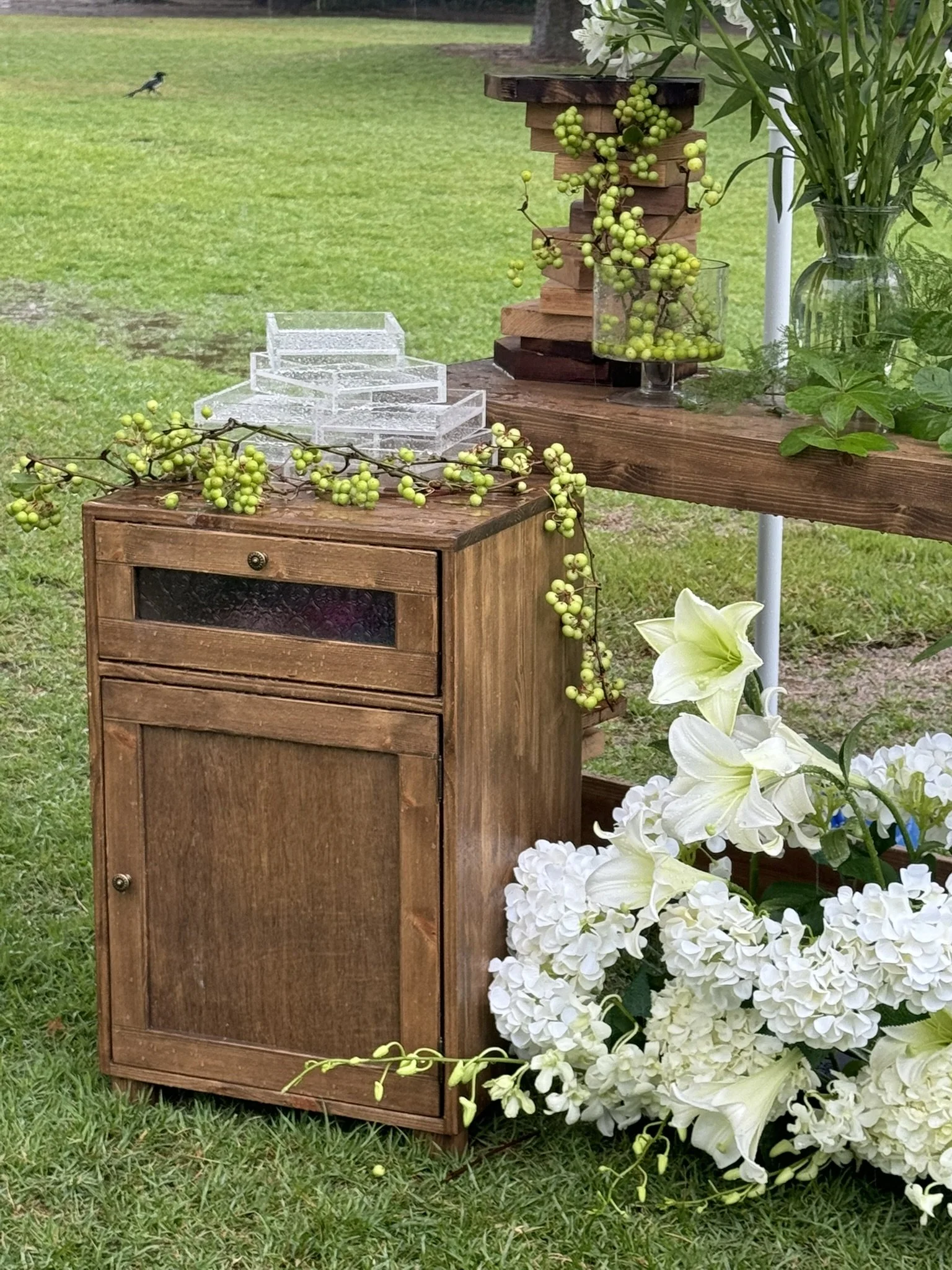 Decorative outdoor setting with a wooden cabinet, green berries, white lilies, and hydrangeas, on a grassy lawn.