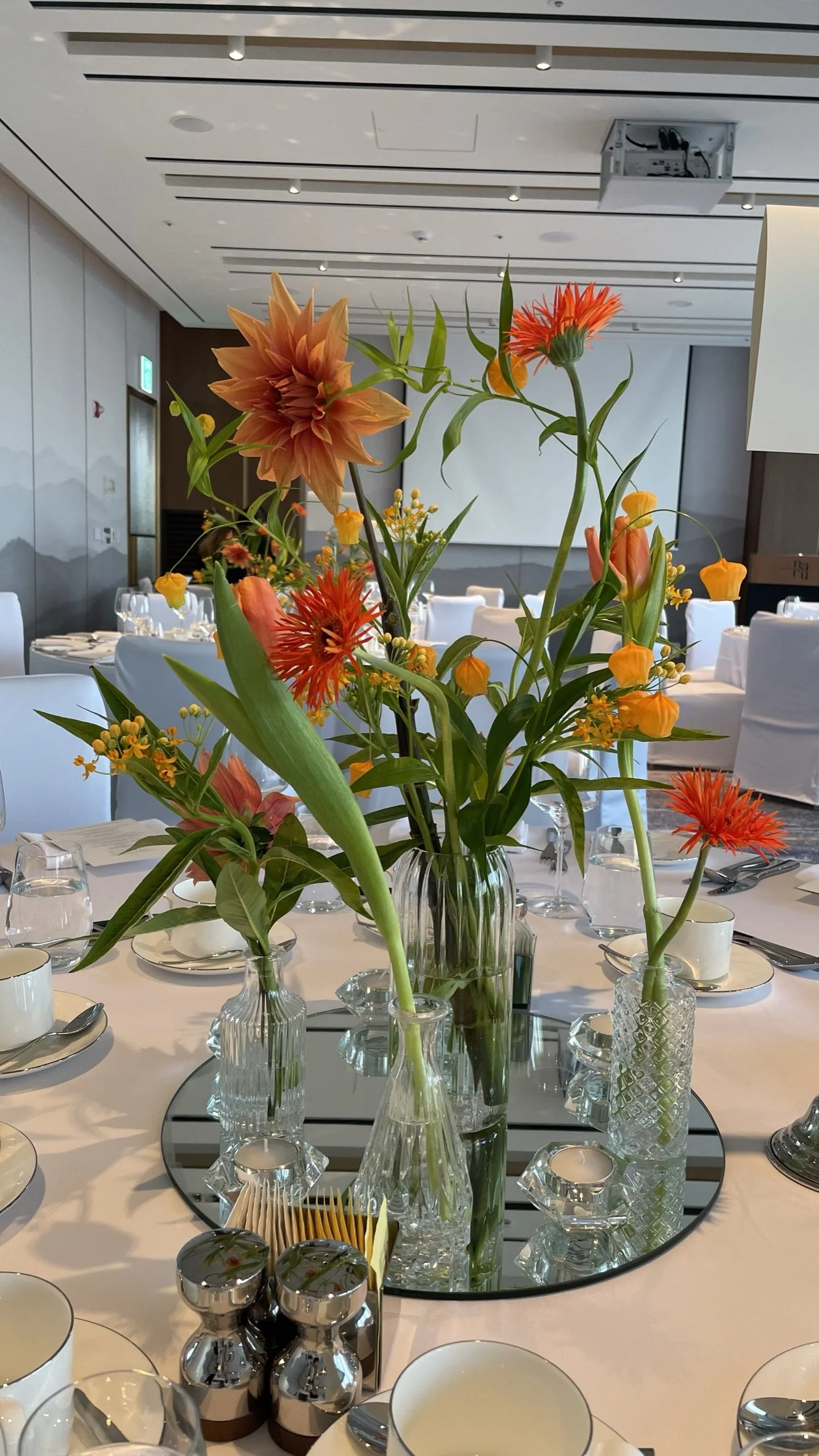 A centerpiece of orange and pink flowers arranged in multiple clear glass vases on a mirror on a white table in a modern event space.