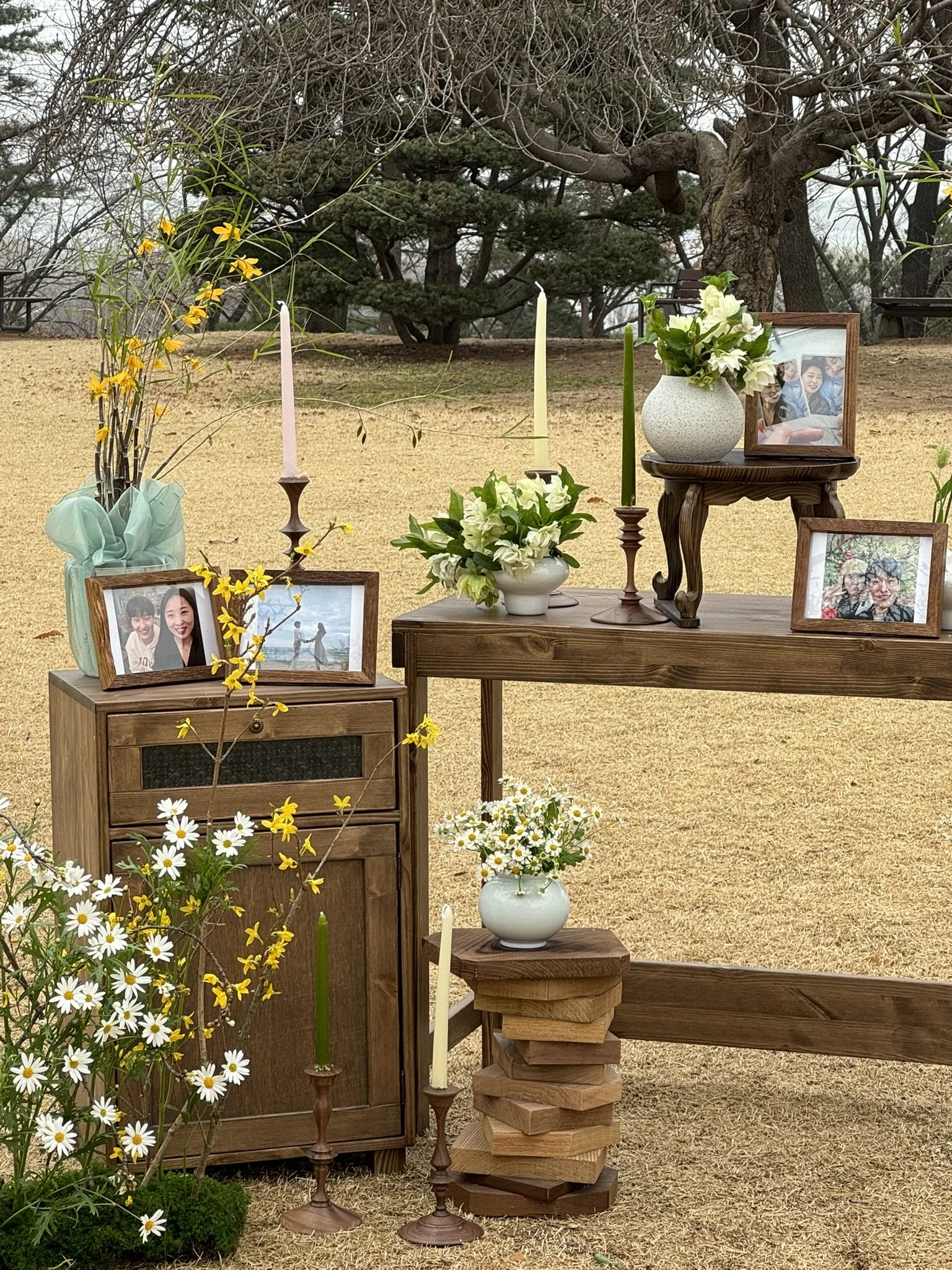 Outdoor memorial setup with framed photos, vases of flowers, candles, and candles on a dirt ground with trees in the background.