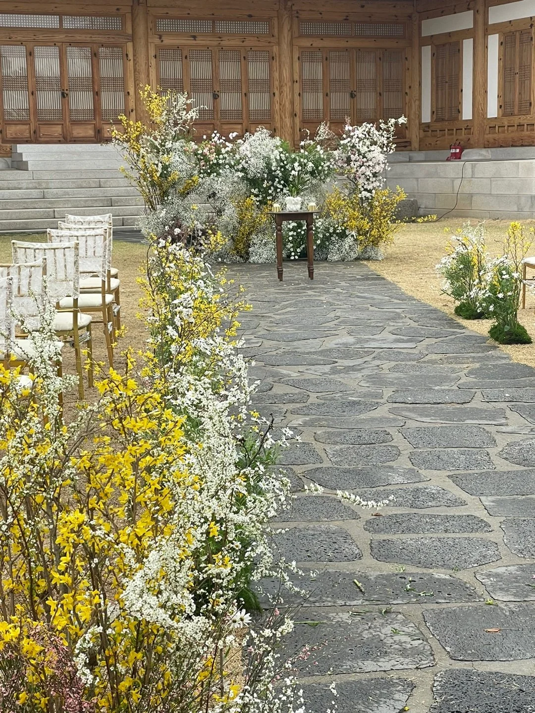 A wedding ceremony setup outdoors with a stone pathway lined with yellow and white flowers leading to a small table at the front, against a wooden building backdrop.