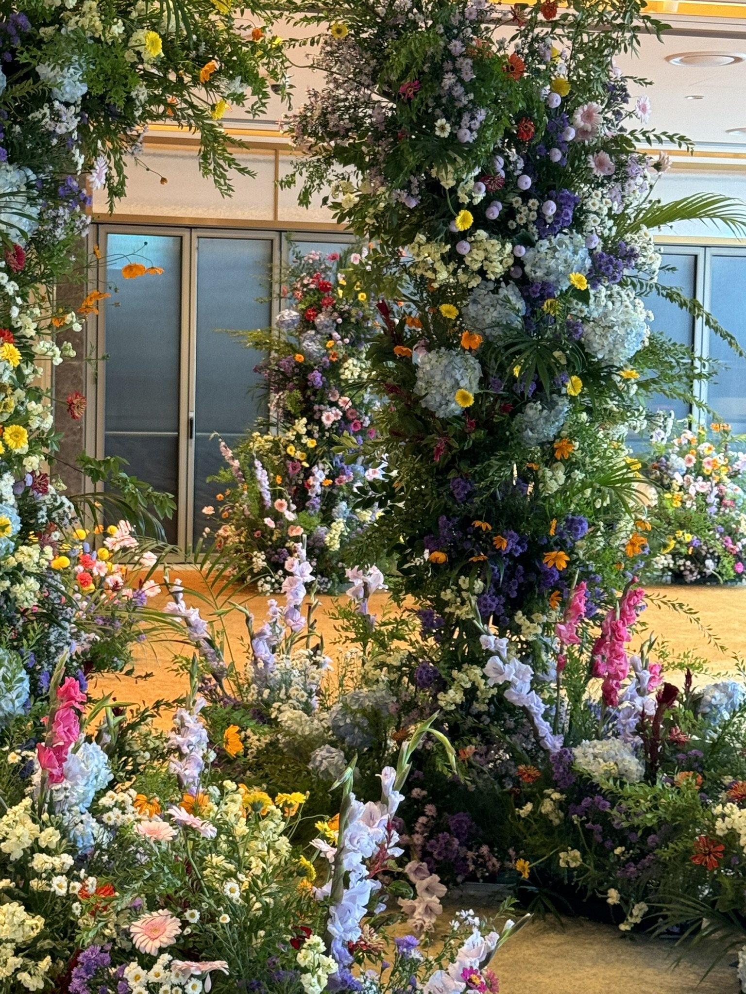 A floral floral archway with a variety of colorful flowers including white, purple, pink, yellow, and orange blooms, set in an indoor space with sliding glass doors and a wooden floor.
