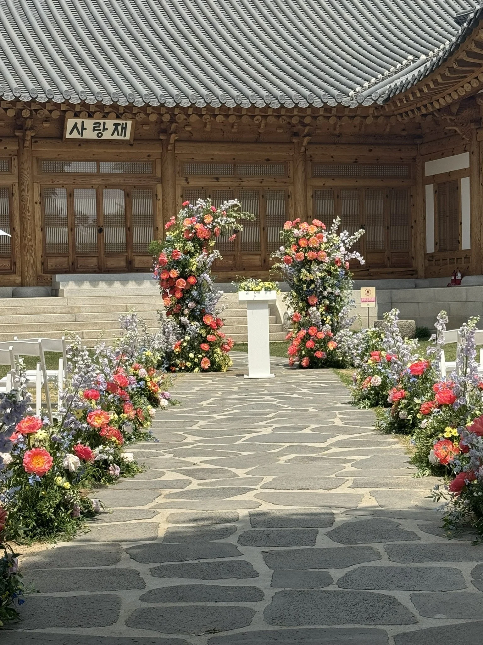 Flowered aisle leading to a wooden building with steps, sign with Korean writing, and blooming trees.