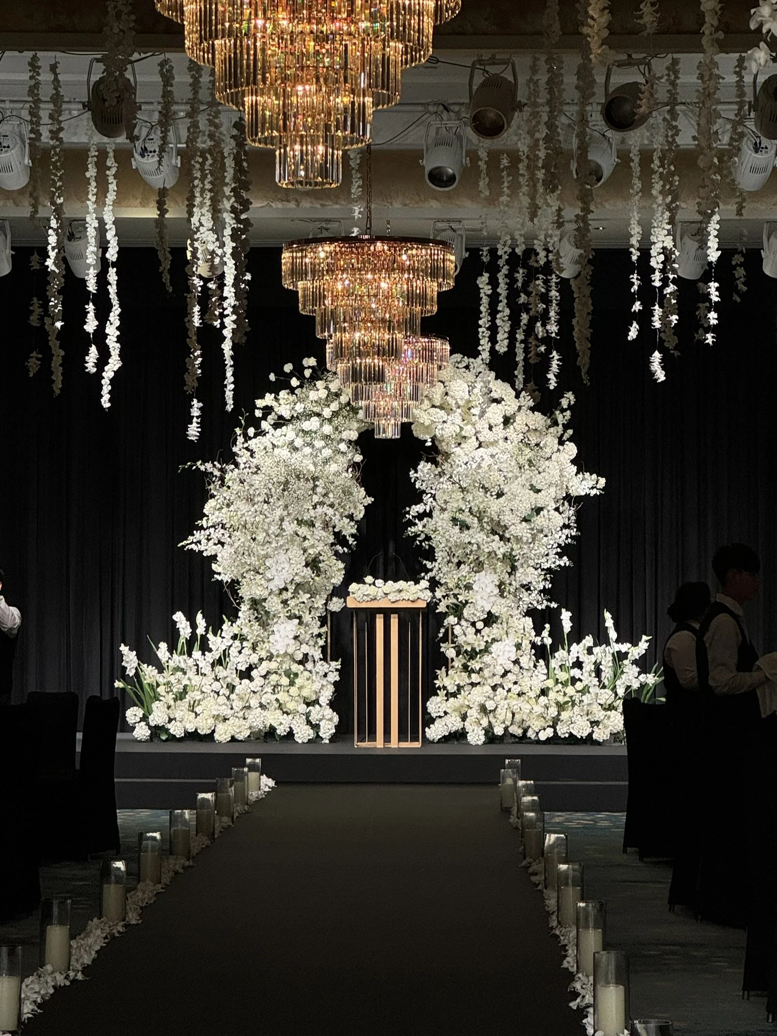 Elegant wedding ceremony setup with a floral arch made of white flowers and greenery, a small podium at the center, and a dark backdrop with hanging white flowers and chandeliers.