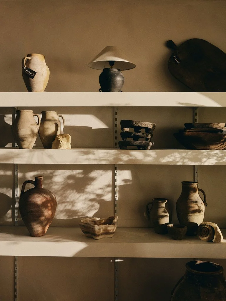 Aardewerken in natuurlijke tinten, zorgvuldig gestyled op witte planken met zacht vallende schaduwen van bladeren.