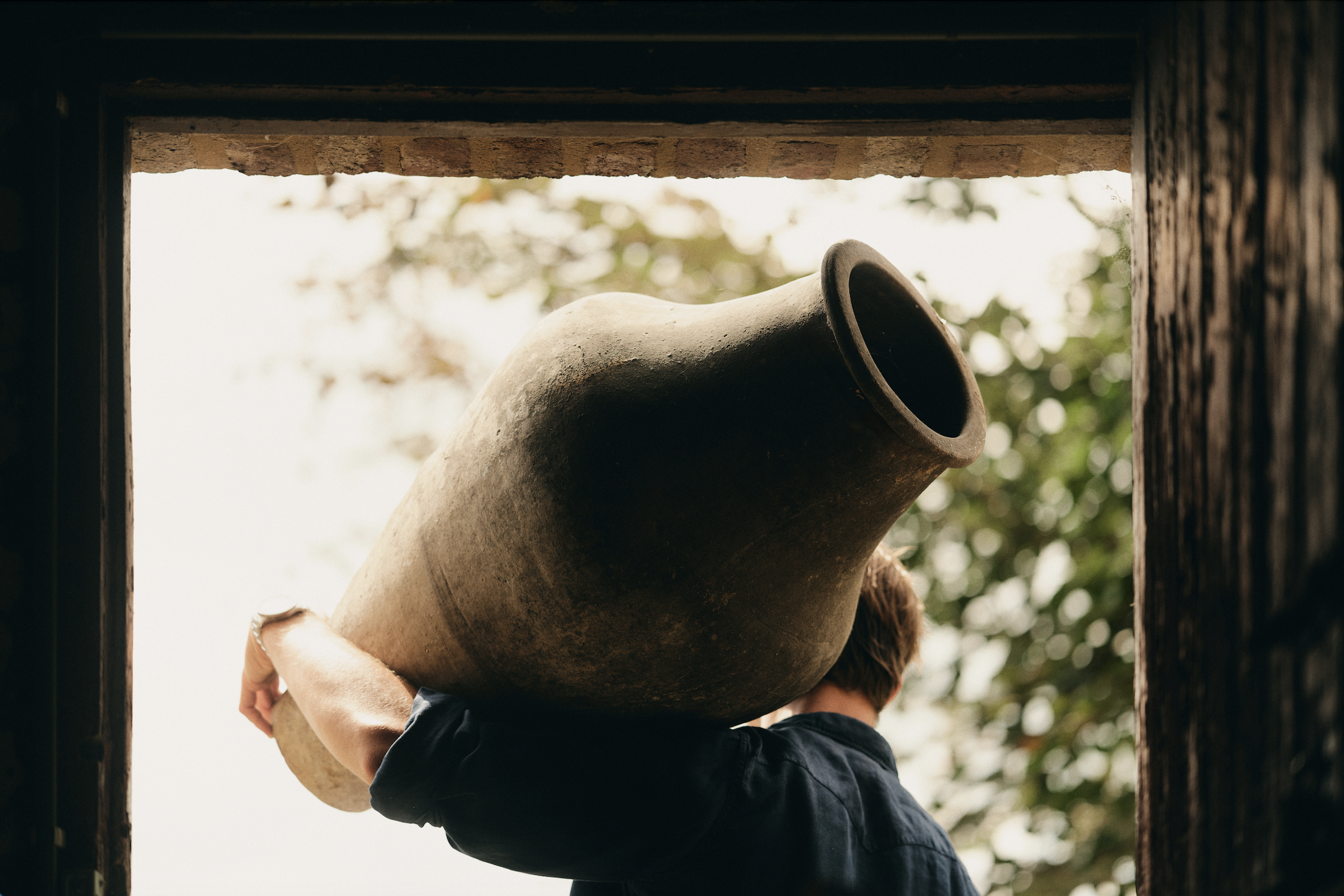 Grote antieke aardewerken pot bij deuropening met bomen op de achtergrond.