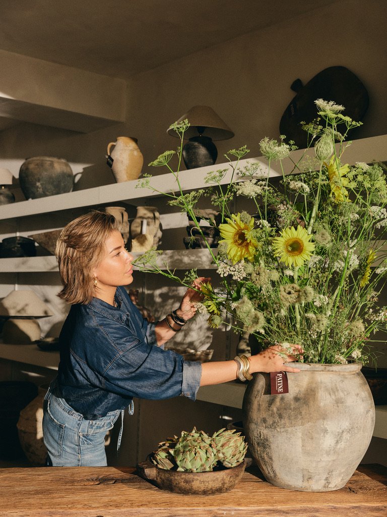 Vrouw schikt boeket in een grote vaas tussen planken met aardewerken potten en vazen