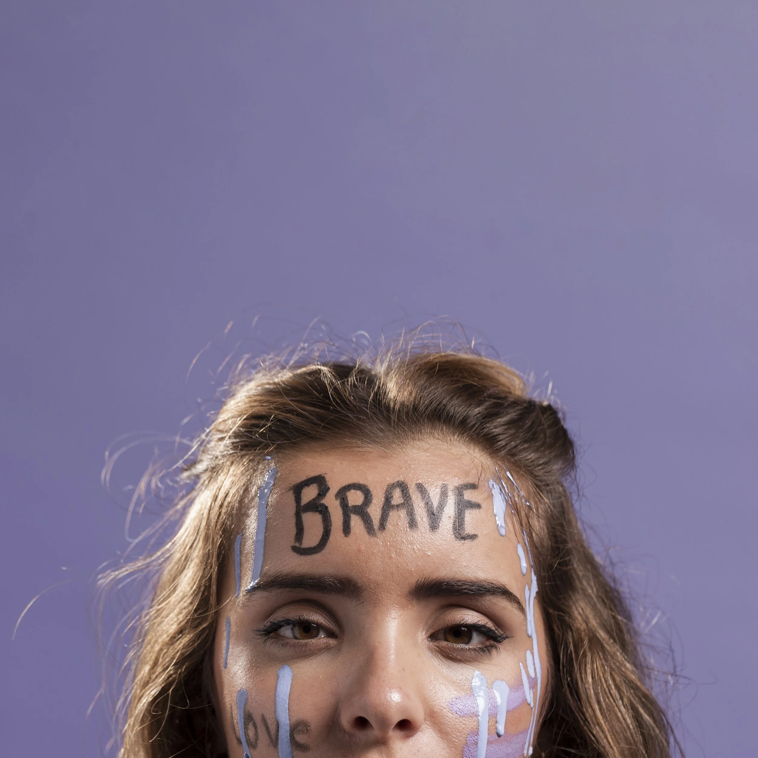 Close up of a woman with the word "BRAVE" written across her forehead and paint streaks on her face, representing reclaiming identity and self worth after relational trauma and emotional abuse.