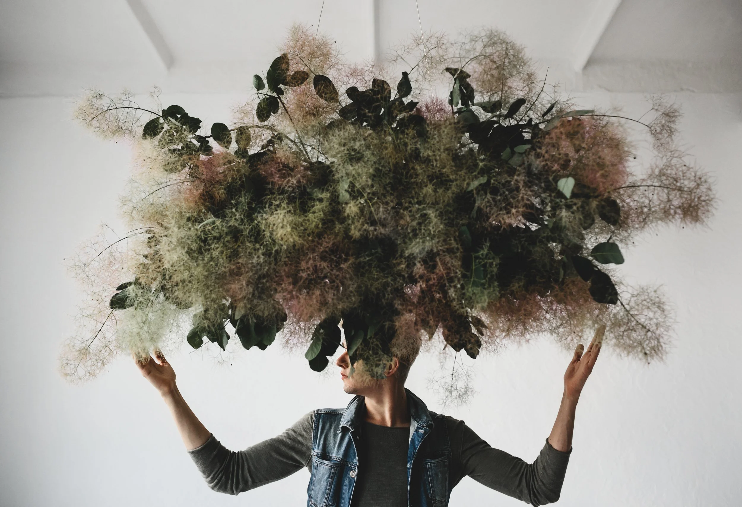 A person stands indoors with arms raised beneath a large cloud of tangled foliage hovering over their head, symbolizing the mental overwhelm and confusion often experienced in relational trauma and trauma bonding.