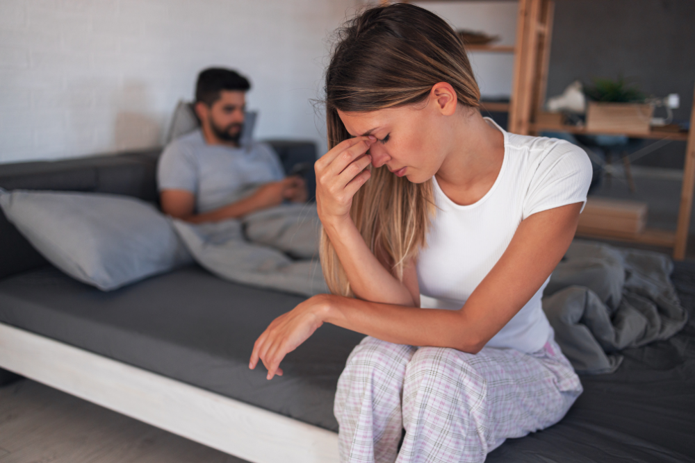 Distressed woman sitting on the edge of a bed wiping tears while her partner sits behind her, depicting emotional abuse in a relationship