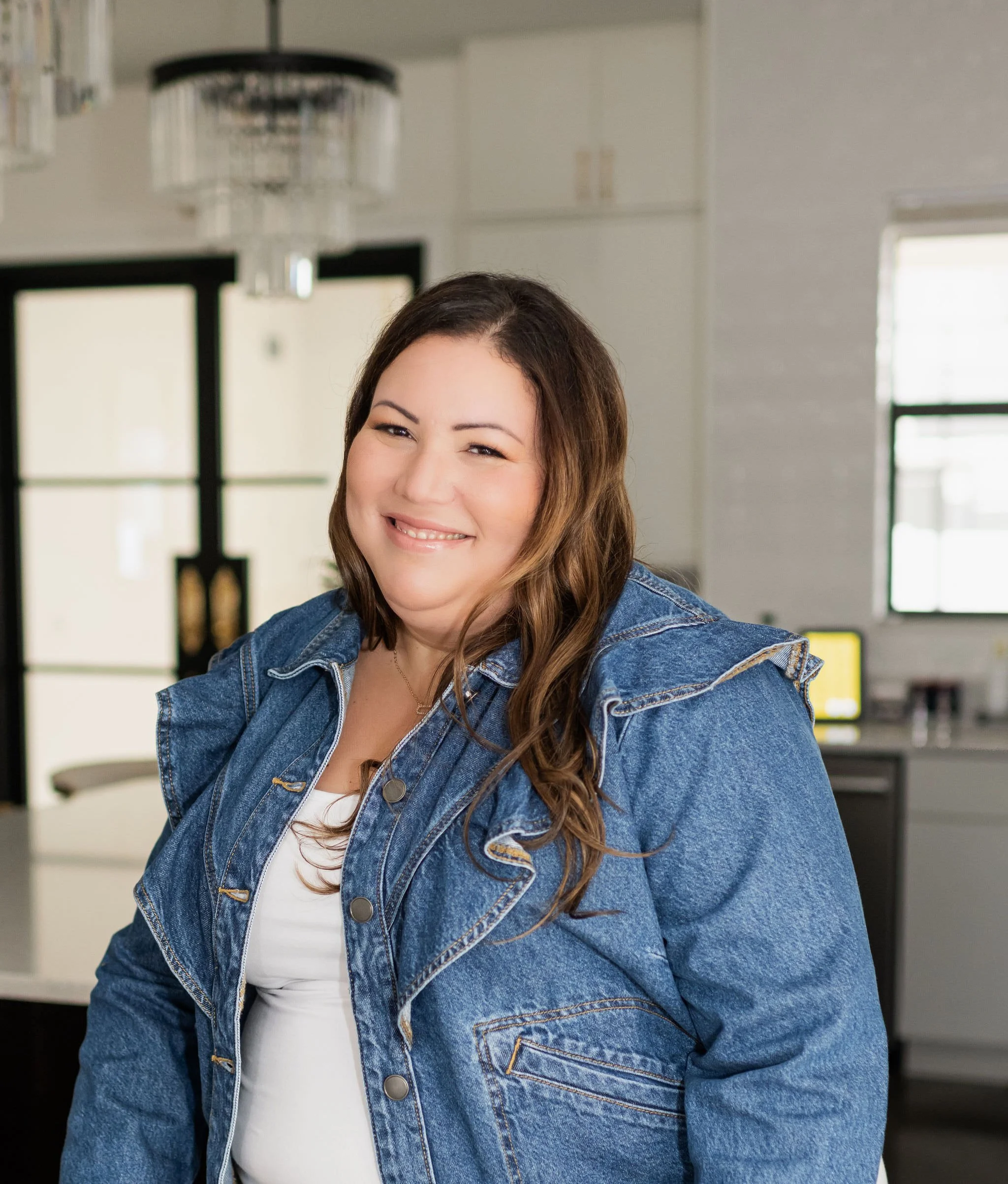 Dr. Katia Arroyo smiling and seated indoors, wearing a denim jacket in a bright, modern setting.