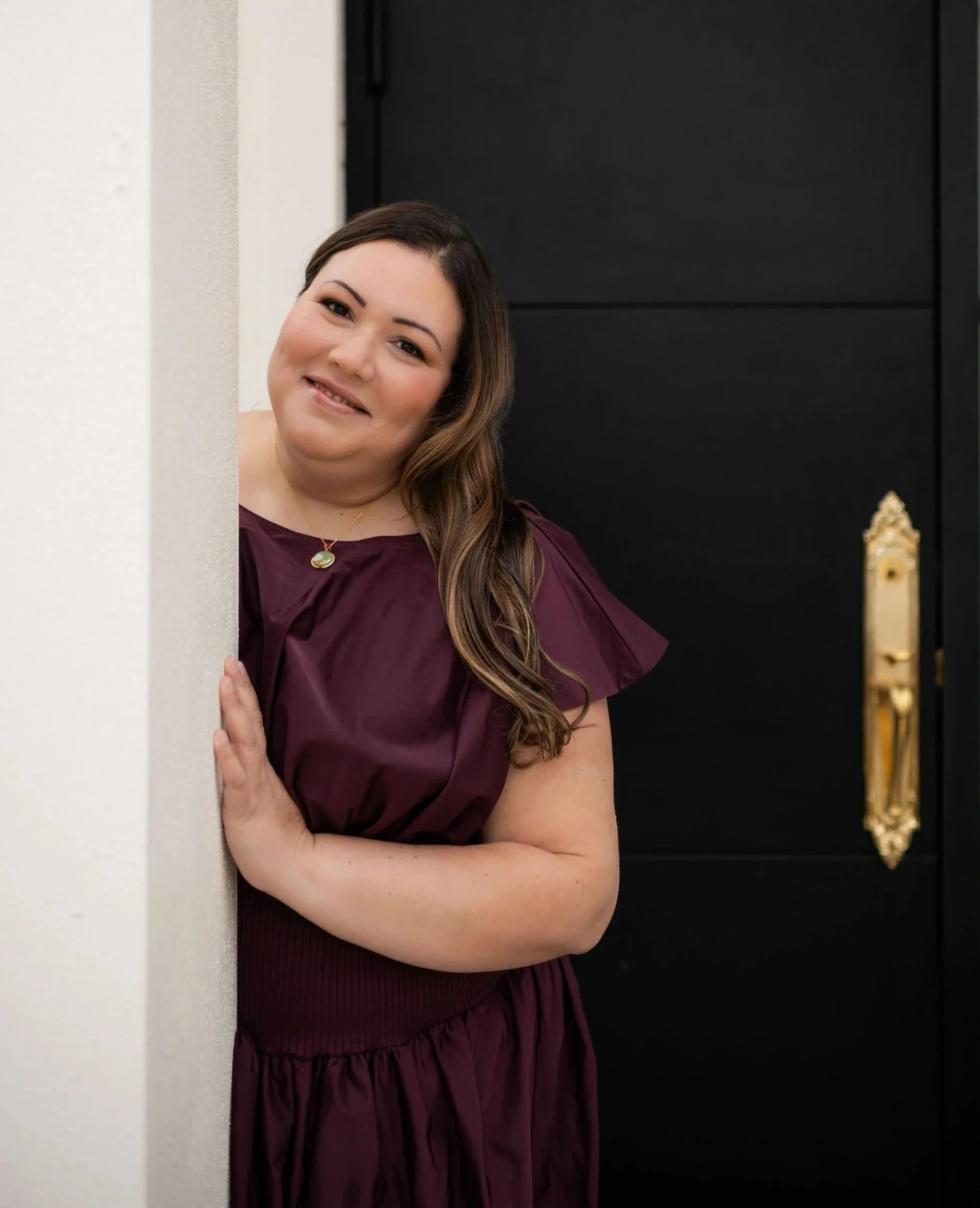 Dr. Katia Arroyo wearing a burgundy dress, standing in front of a black door and peeking from behind a beige wall.