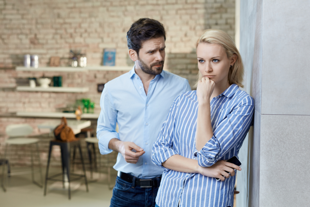 A woman leans against a wall looking distressed while a man stands behind her mid conversation in a kitchen setting, illustrating emotional tension and relational trauma in a romantic relationship.