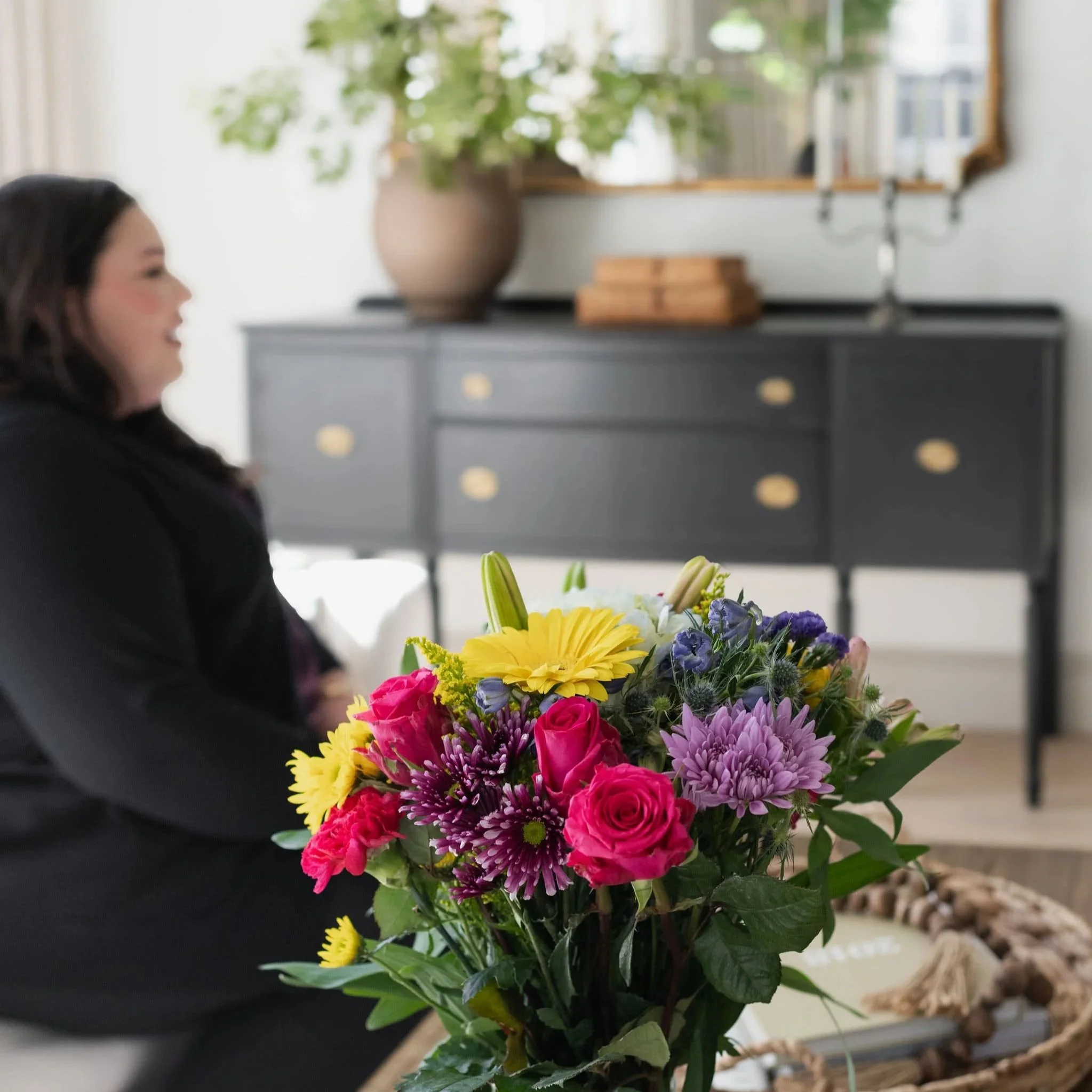 Dr. Katia Arroyo seated indoors in a softly lit room, with a colorful bouquet of flowers in the foreground and a dark console table behind her.