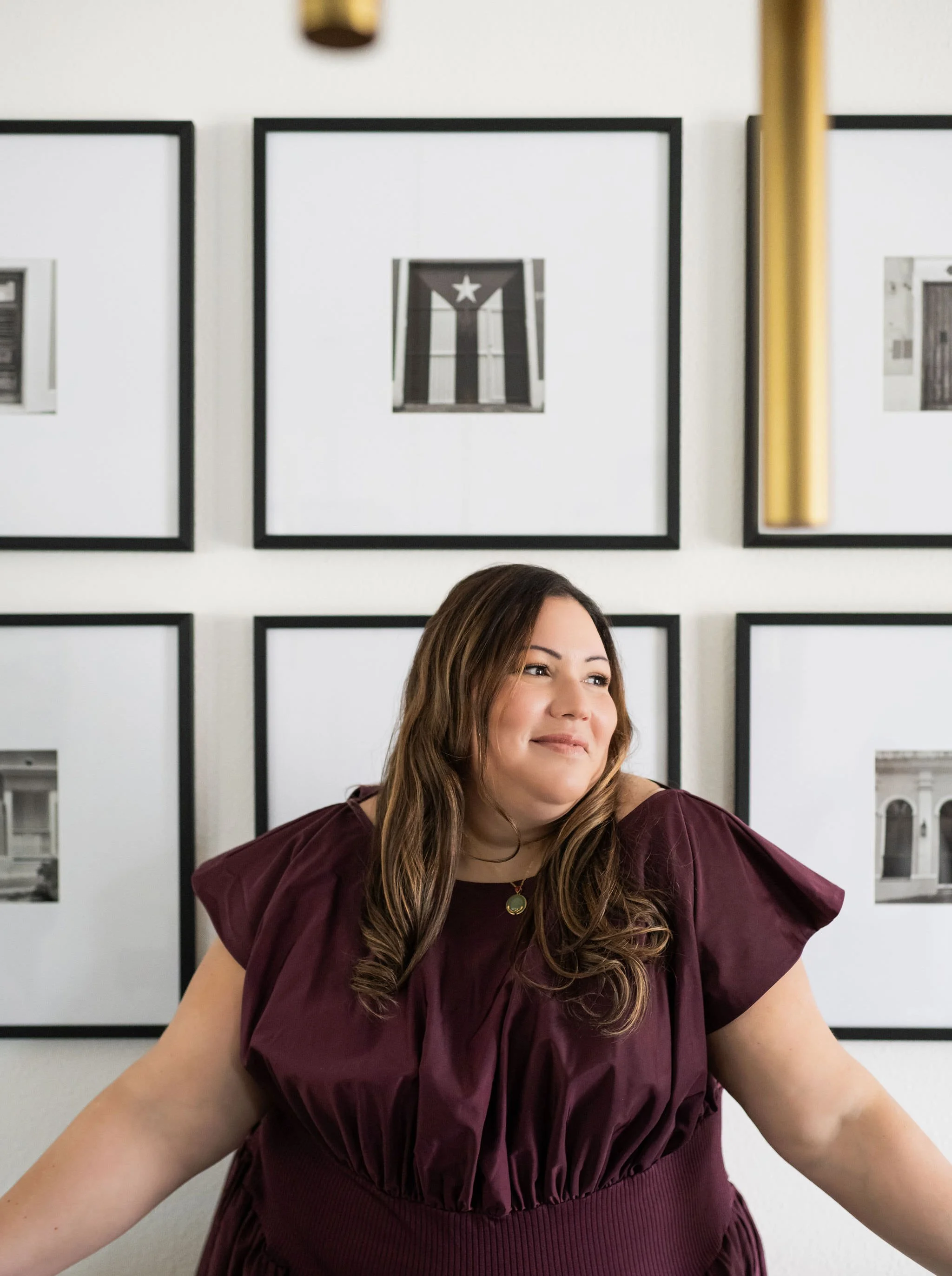 A woman with long wavy brown hair wearing a burgundy dress sitting at a table in front of a gallery wall with framed black-and-white photographs.