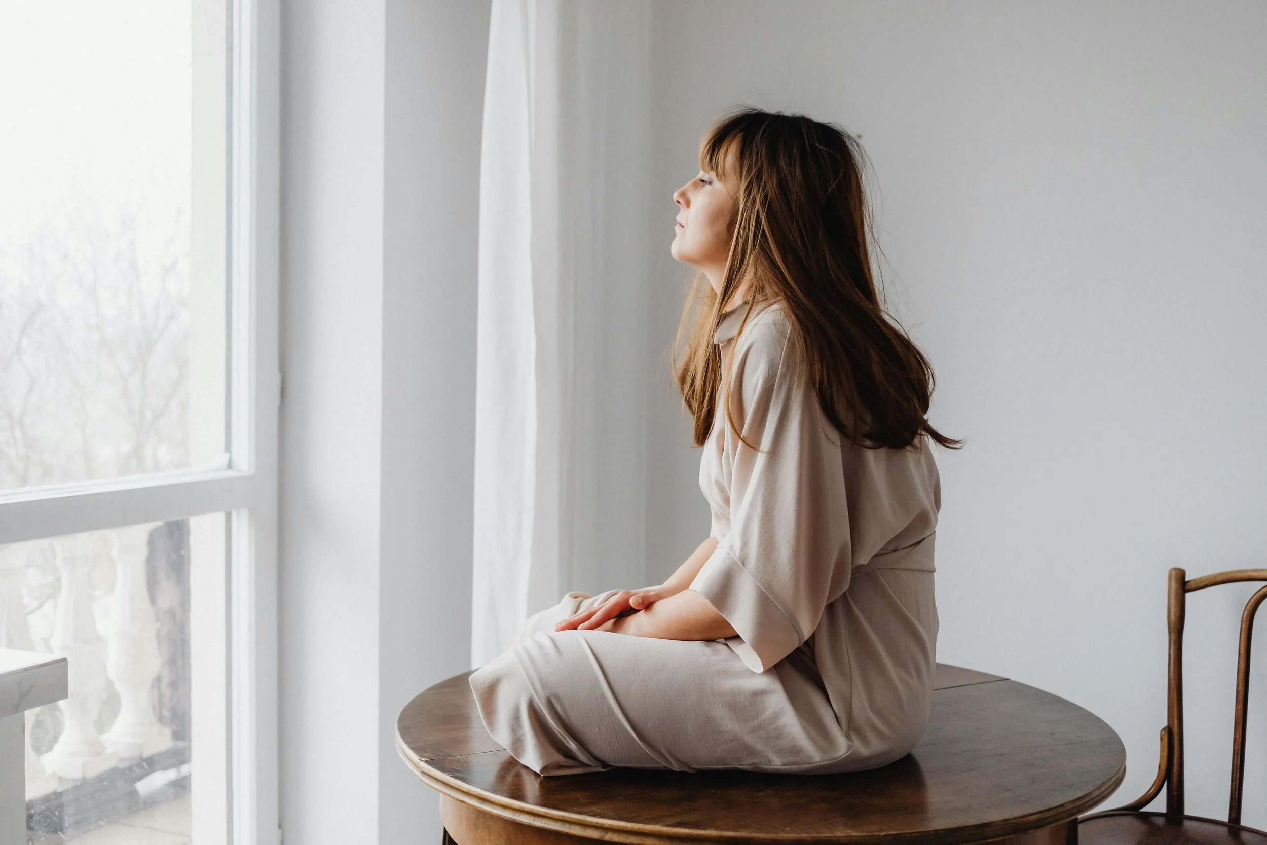 Woman sitting cross-legged on a wooden table by a sunlit window, reflecting on trauma and burnout