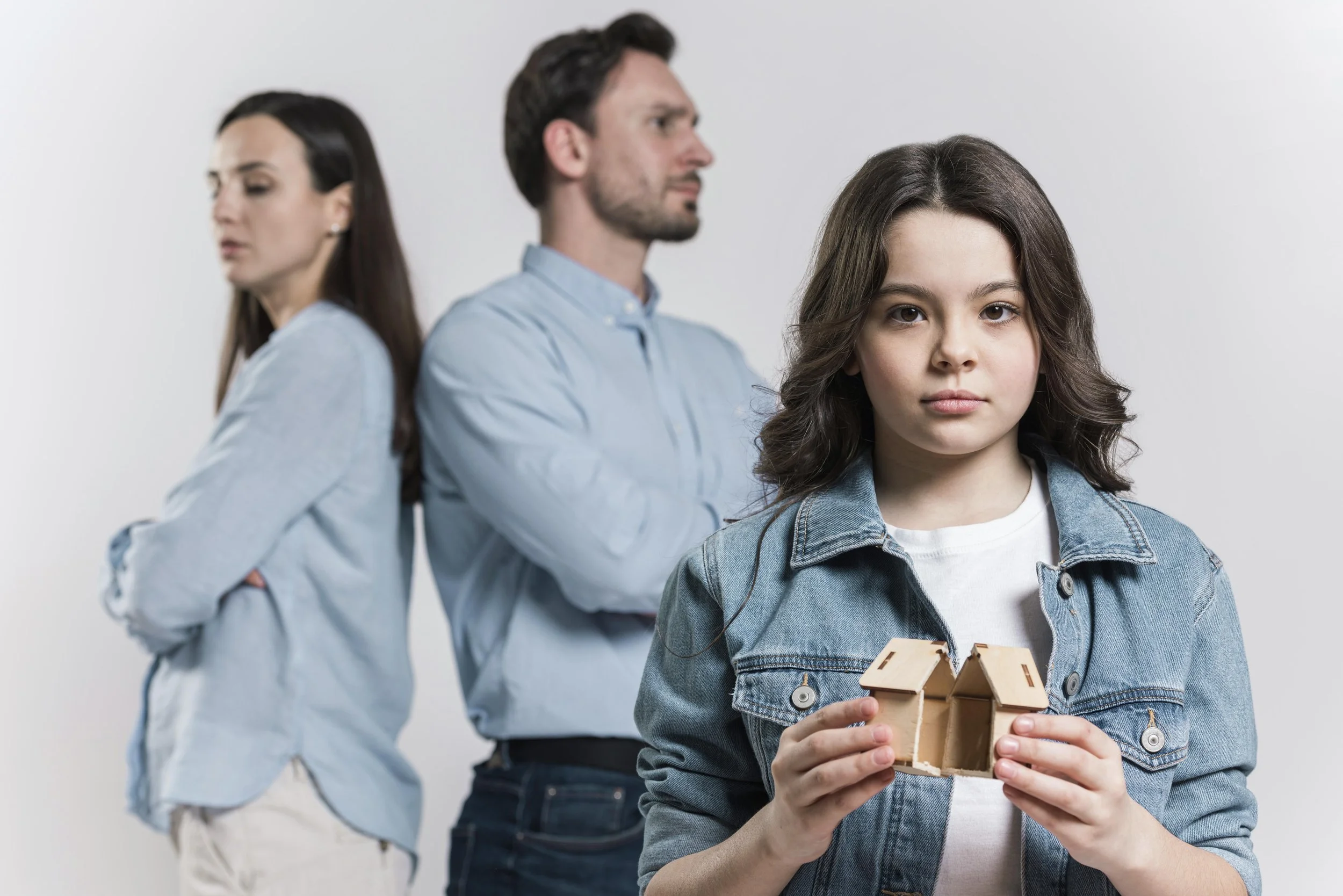 Young girl holding a small wooden house looks toward the camera while her parents stand behind her turned away from each other, showing tension in the home and the impact of generational trauma on children.
