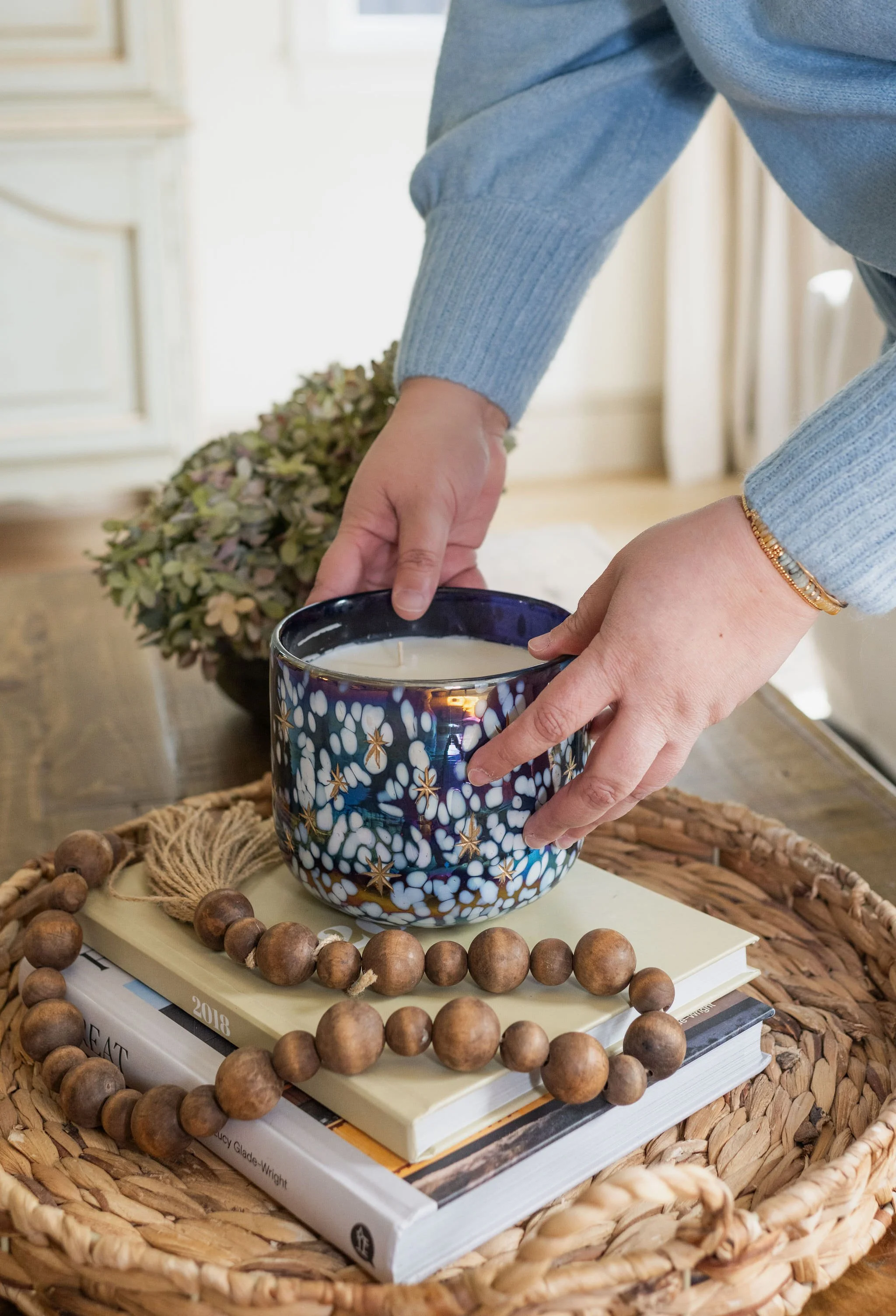 Hands placing a decorative blue candle on top of stacked books on a woven tray.