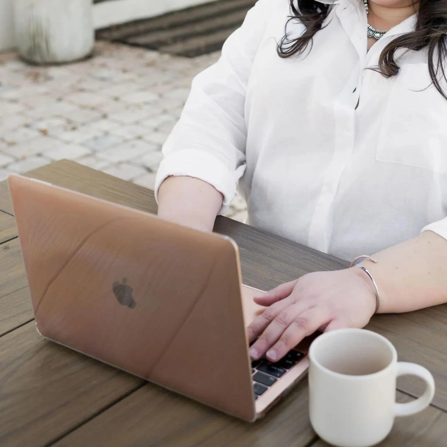 Person in a white blouse typing on a laptop at an outdoor wooden table with a coffee mug nearby.