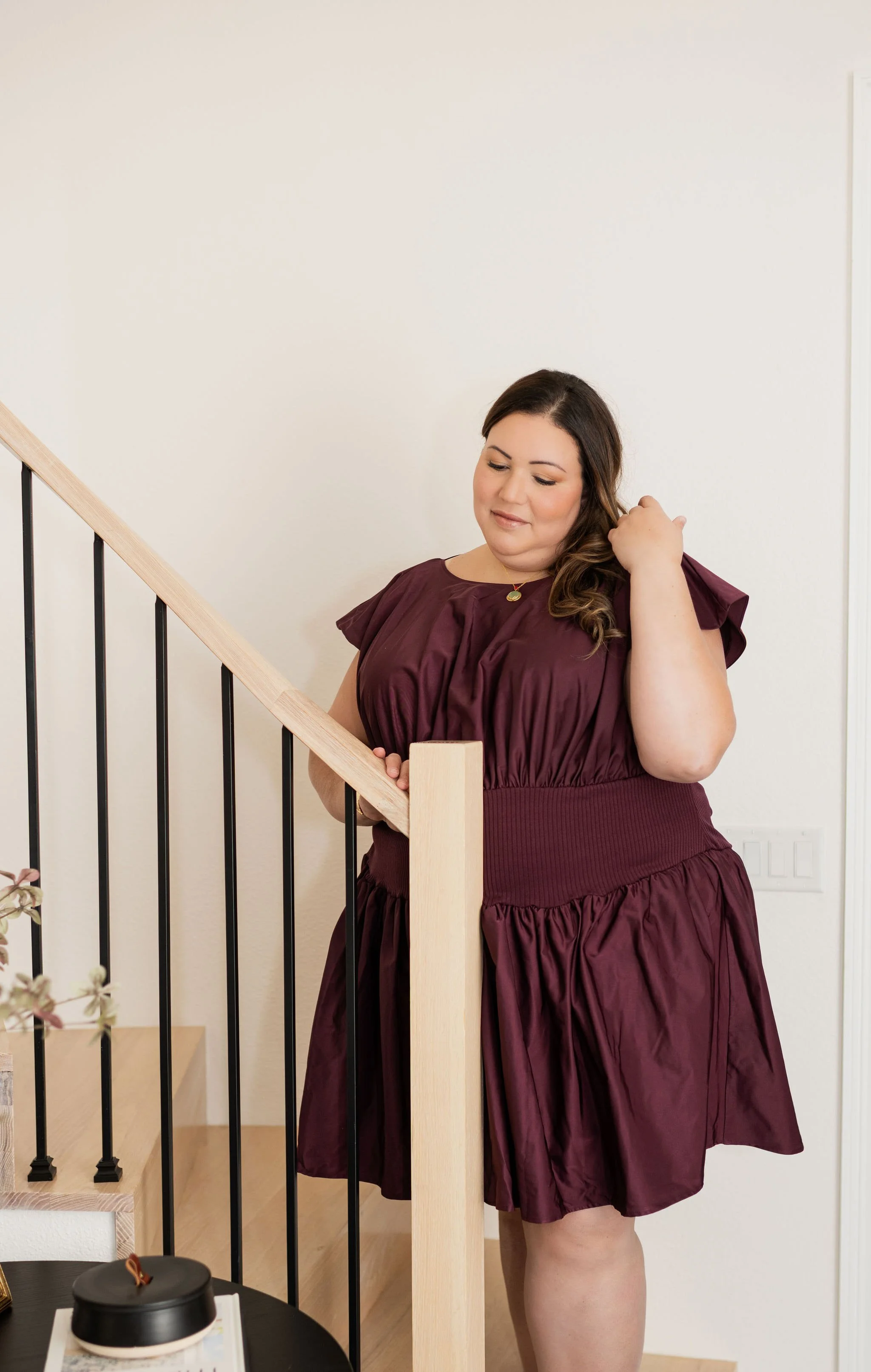 Dr. Katia Arroyo wearing a burgundy dress, standing on a staircase and gently adjusting her hair.