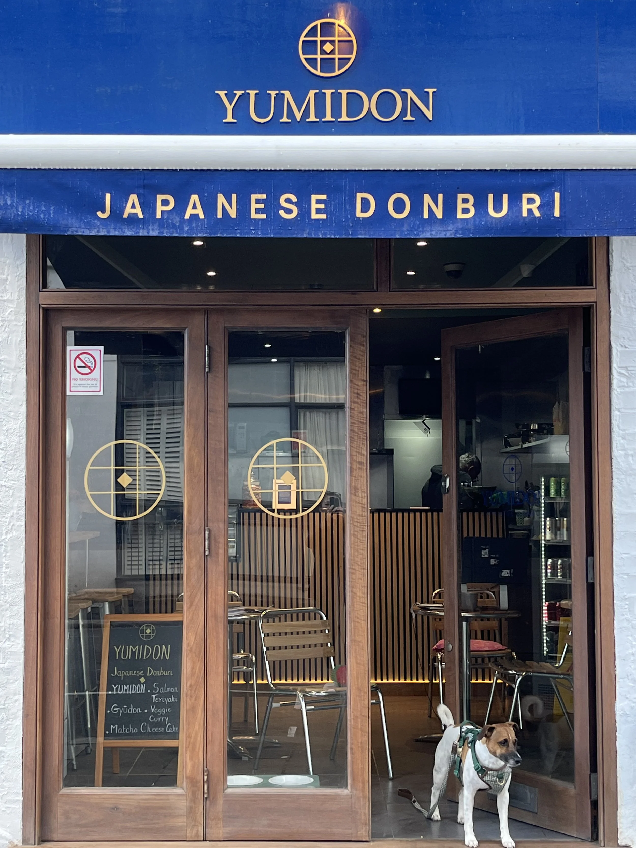The exterior of a Japanese donburi restaurant called YUMIDON, with wooden framed glass doors, a blue and gold sign, and a dog standing outside.