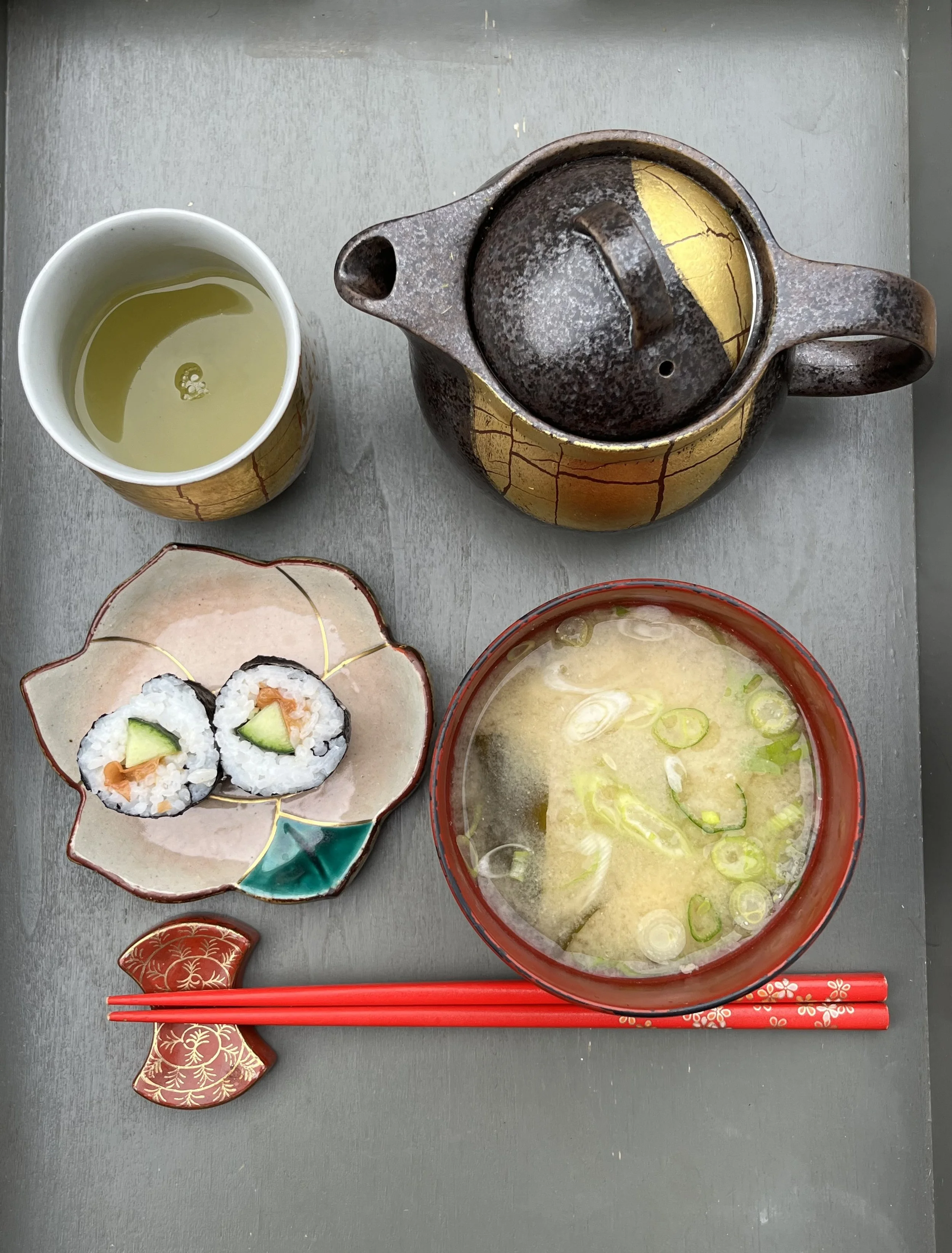 A Japanese-style meal on a gray table, featuring a bowl of miso soup with green onions, two sushi rolls with cucumber and salmon, a cup of green tea, a textured teapot with a yellow and black pattern, and red chopsticks with a decorative fish-shaped rest.