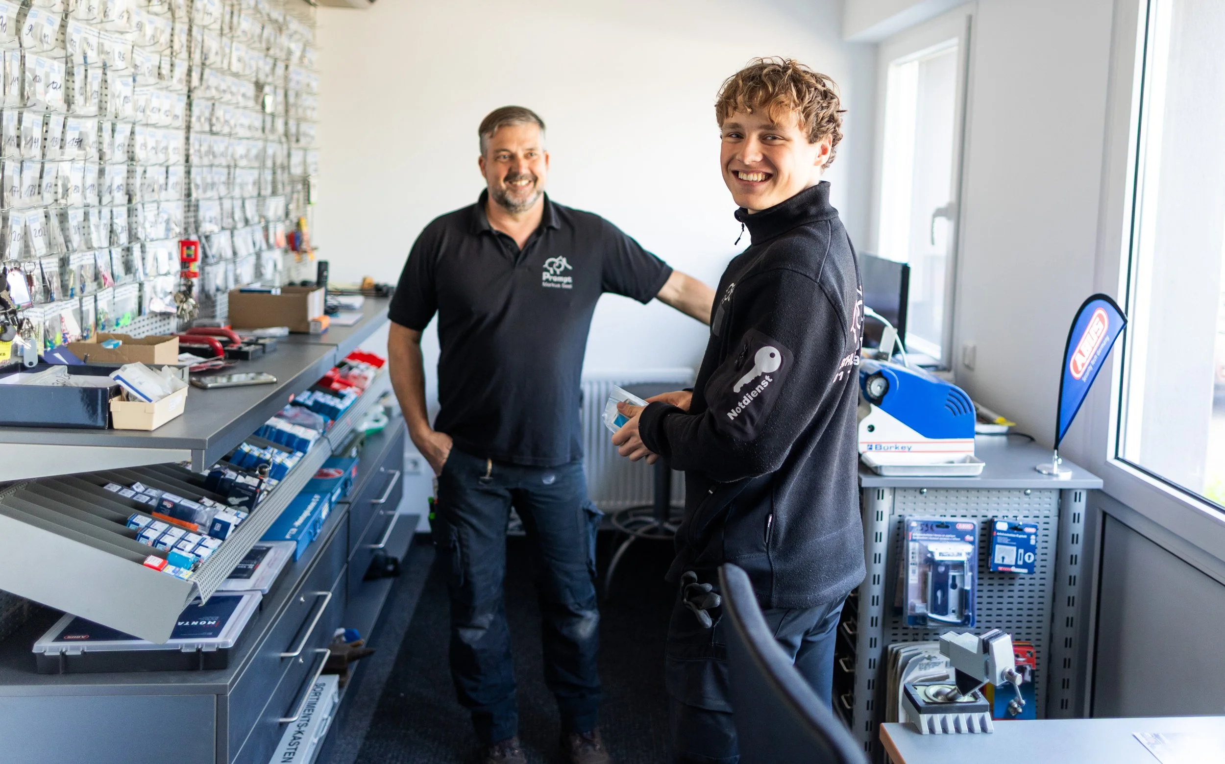 Zwei Männer in einem Werkstatt- oder Lagerraum, die freundlich lächeln. Der ältere Mann trägt ein schwarzes Polo-Shirt, der jüngere Mann trägt eine schwarze Jacke mit Logos. Der Raum hat Regale mit kleinen Teilen und Werkzeugen.